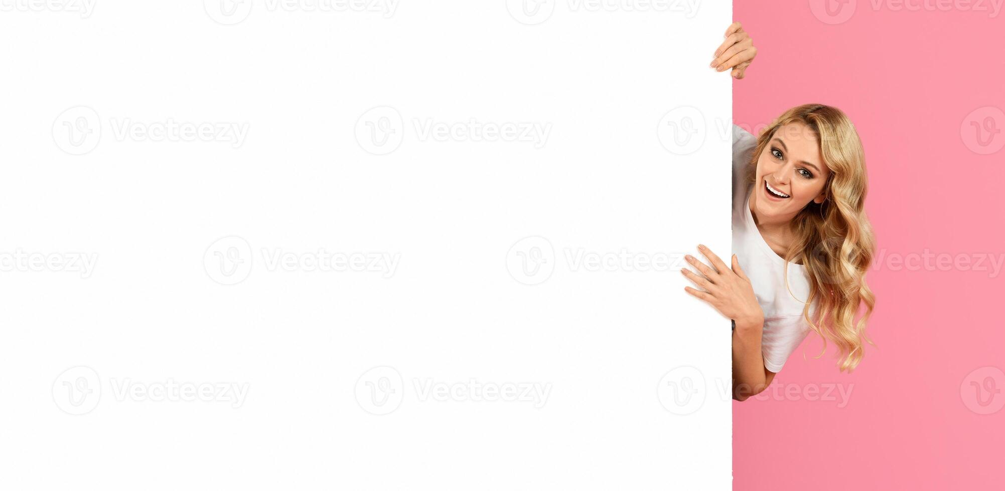 A cheerful young woman with long blonde hair playfully peers from behind a blank white board against a vibrant pink backdrop. She exudes excitement and positivity in this lively setting. photo