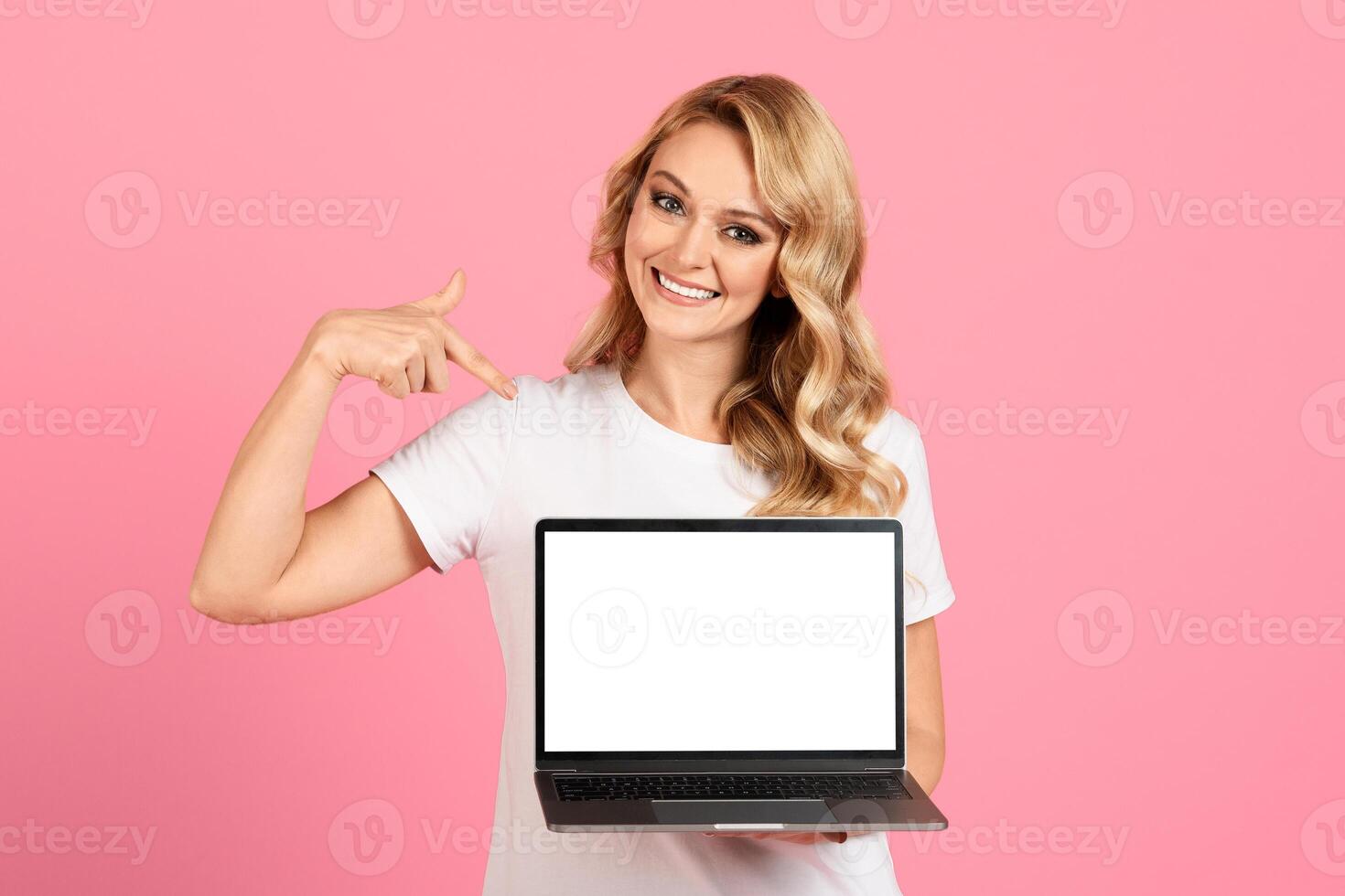 A cheerful young woman points to a blank laptop screen while standing in front of a bright pink background. She has a warm smile and casual attire, inviting engagement and creativity. photo