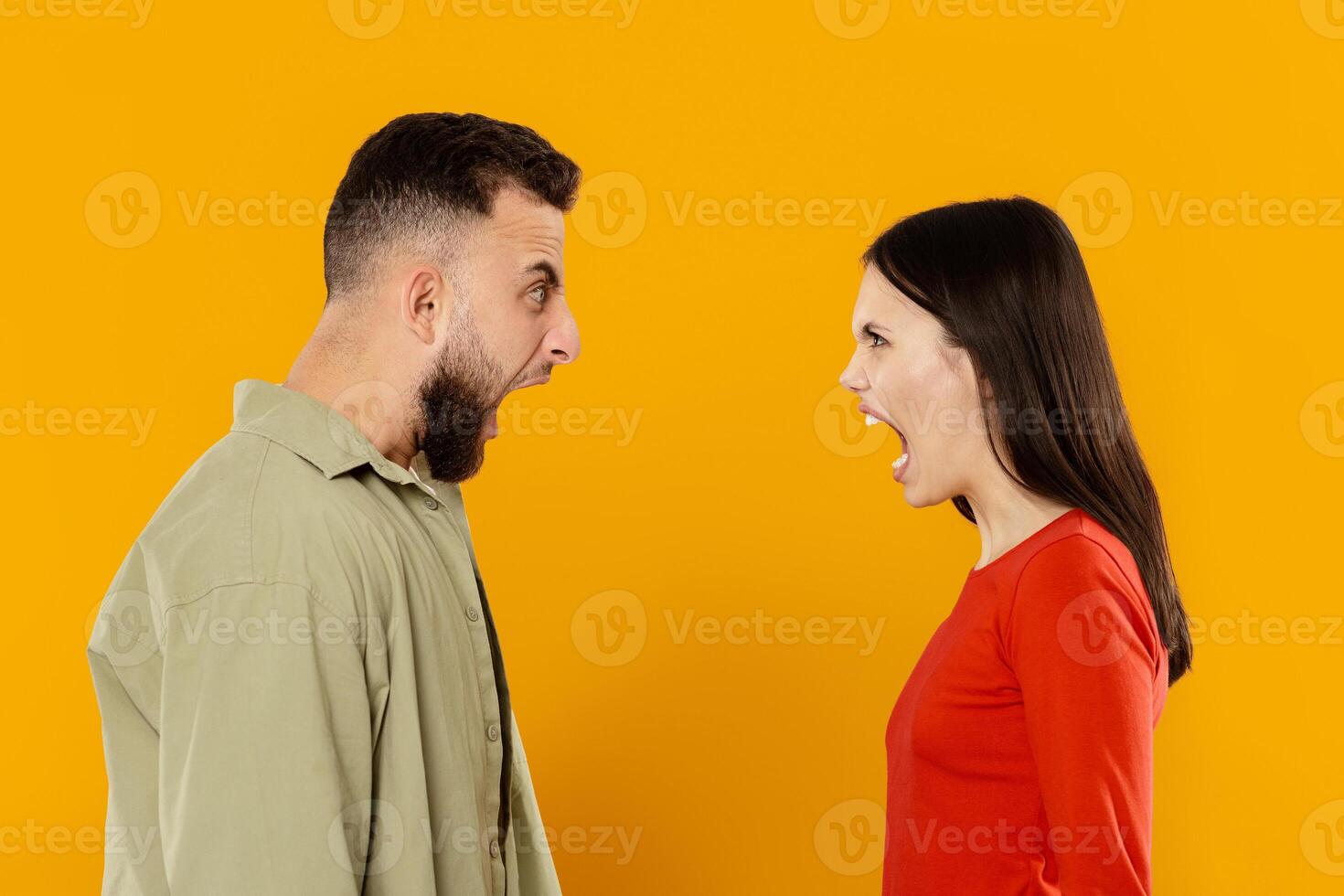 Young couple stands facing each other against a bright yellow background, both expressing intense emotions. They are shouting, showcasing their disagreement and strong feelings. photo