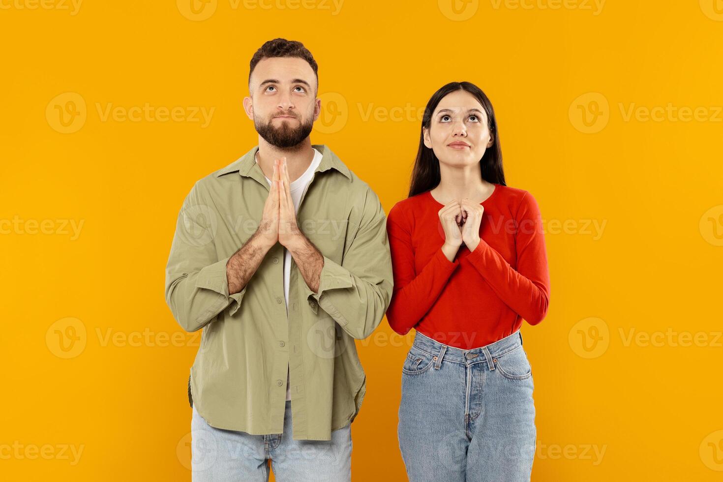 A young couple stands together with hands clasped in anticipation. They look up with hopeful expressions, surrounded by a vibrant yellow backdrop that enhances their joyful connection. photo
