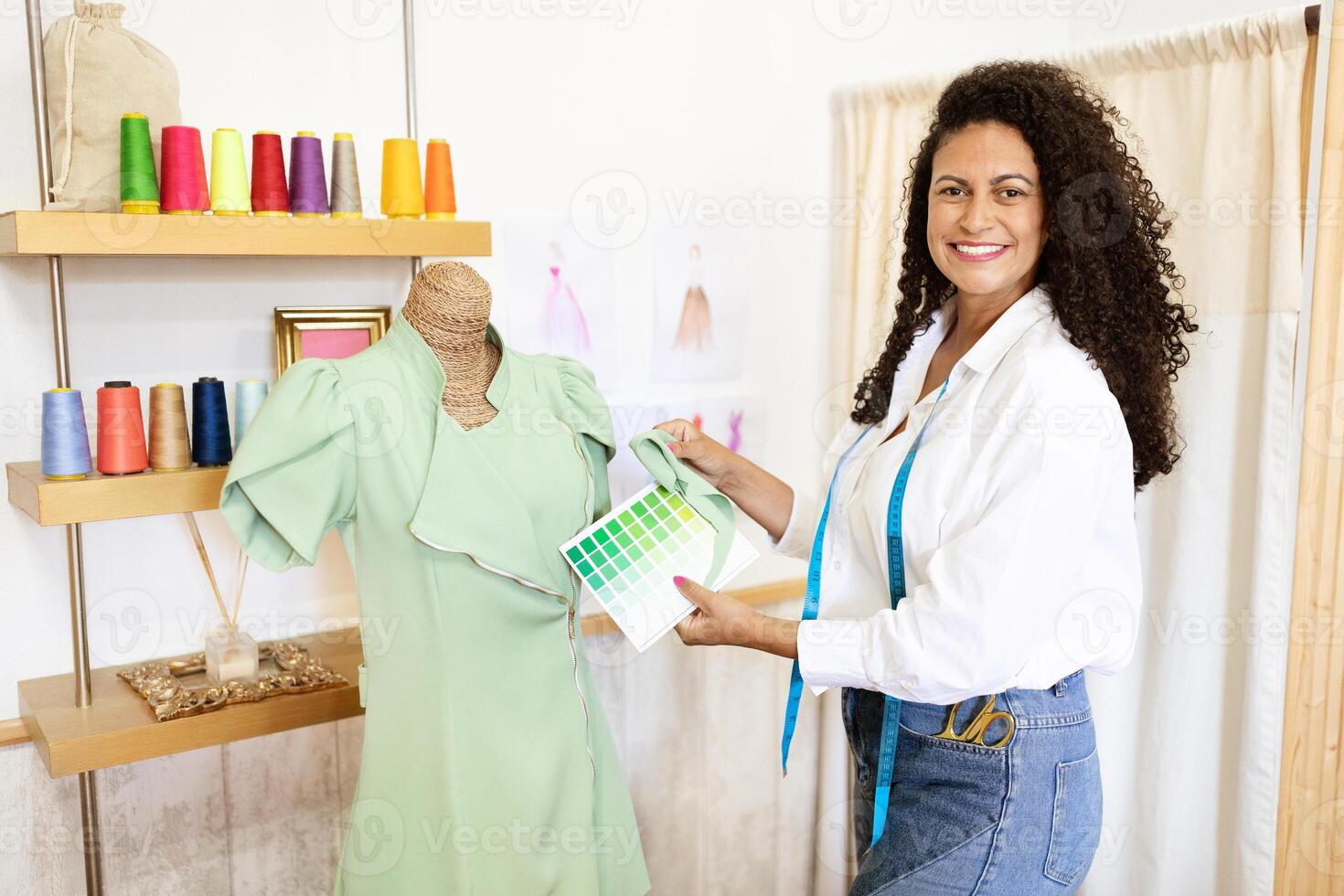 A mature Hispanic woman is smiling while holding a fabric swatch and standing next to a dress on a mannequin. She works in a bright, organized design studio filled with colorful materials. photo