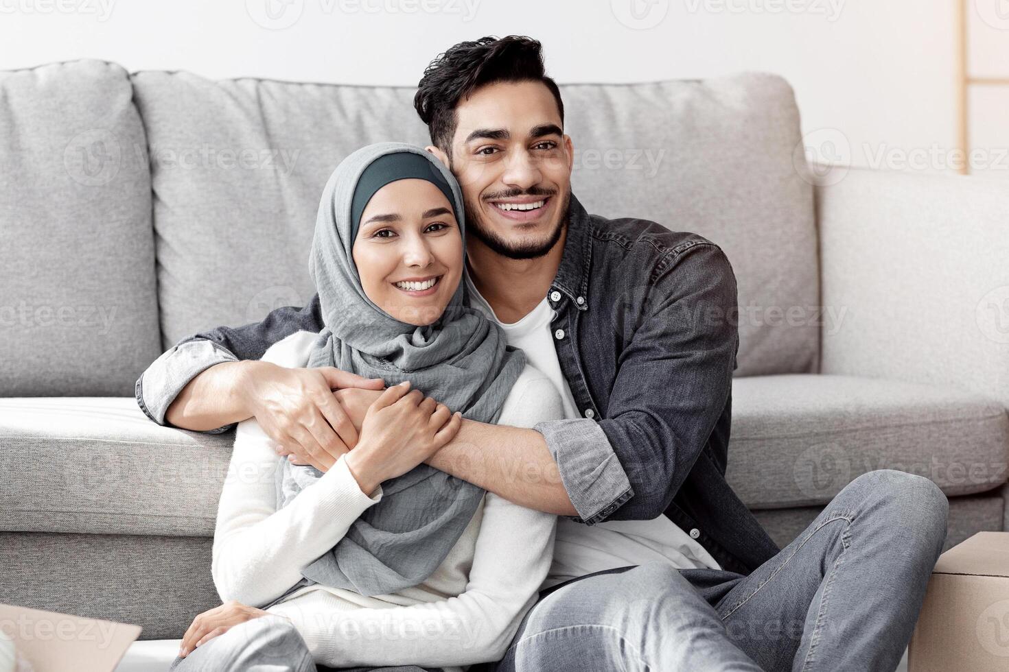 Beautiful middle-eastern couple resting while unpacking after moving house, happy arab guy and lady in hijab sitting on floor by sofa among paper boxes in living room, hugging and smiling, copy space photo