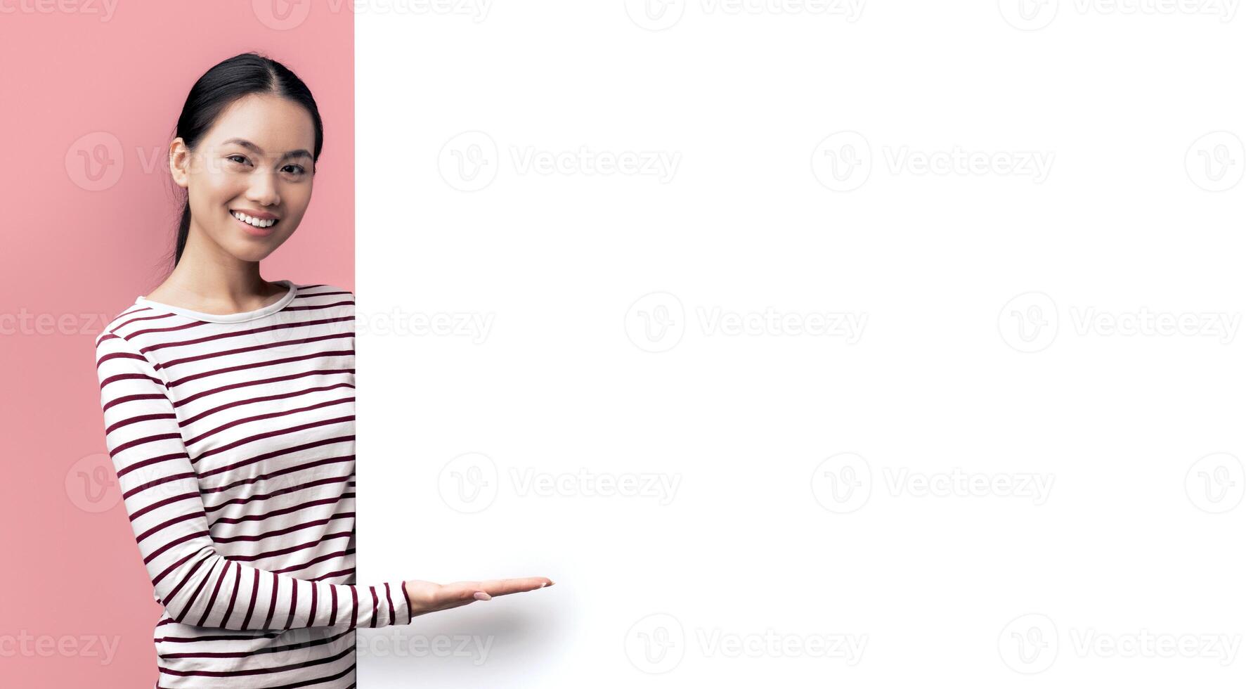 Beautiful Young Smiling Asian Lady Pointing At Big White Advertisement Board With Open Palm, Positive Korean Female Showing Free Copy Space On Blank Billboard, Posing Over Pink Studio Background photo