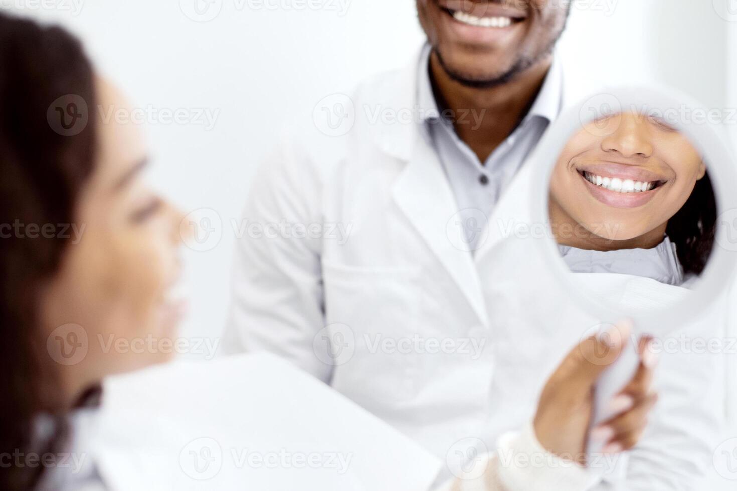 Happy Young Black Woman Looking At Her Teeth In Mirror After Dental Treatment At Stomatological Clinic, African American Female Patient Enjoying Result Of Quality Medical Services, Selective Focus photo