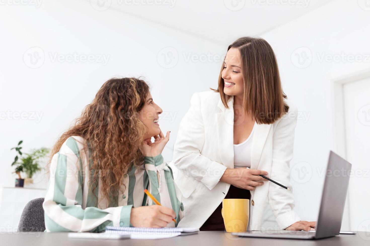 Two European women engage in a collaborative discussion in a bright corporate office. One woman sits with notes, while the other stands and shares insights, fostering teamwork. photo