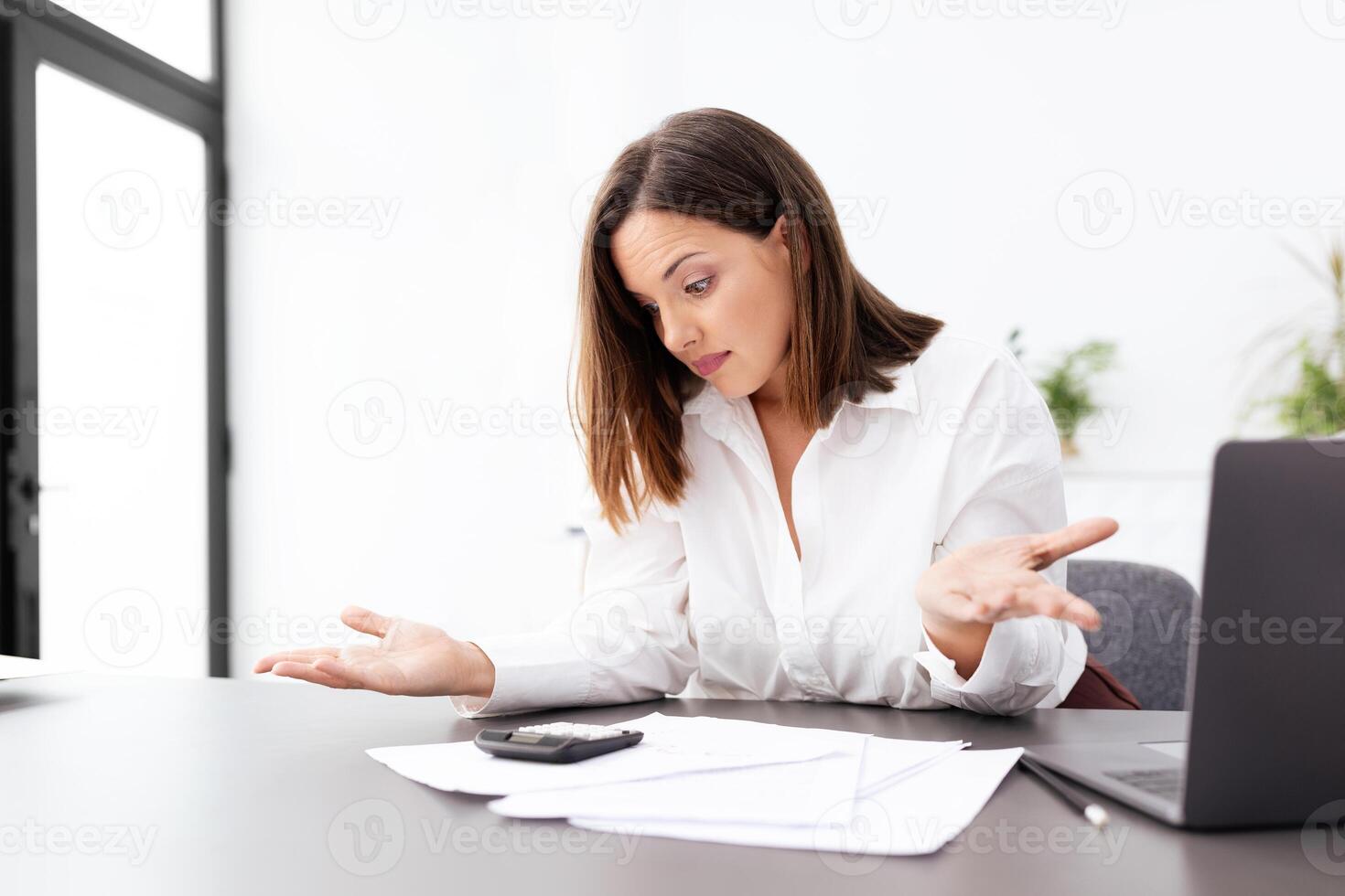 A businesswoman appears frustrated while working at her desk, surrounded by paperwork and a laptop. She gestures with her hands, indicating confusion or stress in a modern office. photo