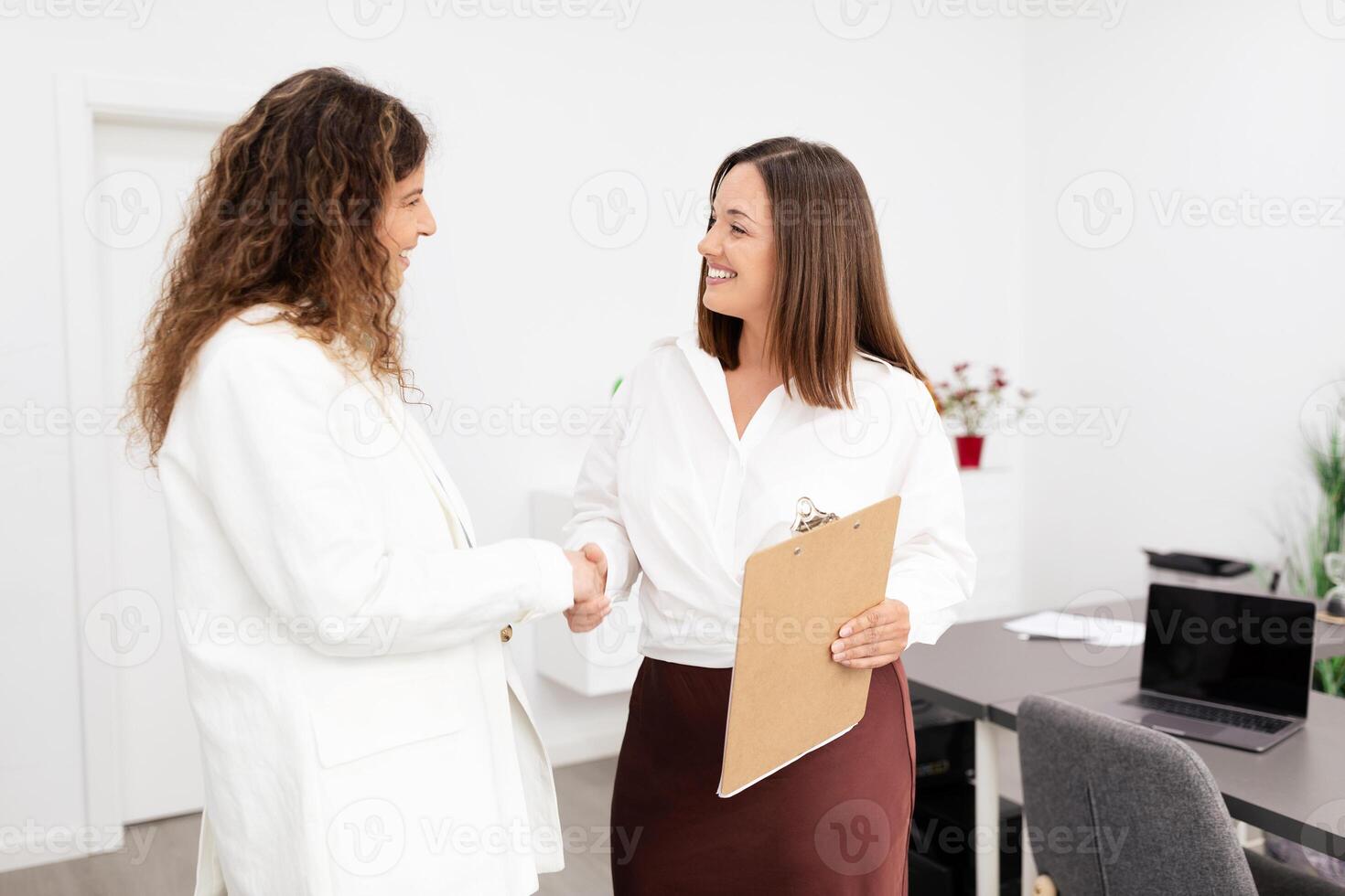 Two Caucasian women engage in a handshake within a modern office environment. One holds a clipboard, indicating a potential collaboration or business meeting. It's a bright, professional space. photo
