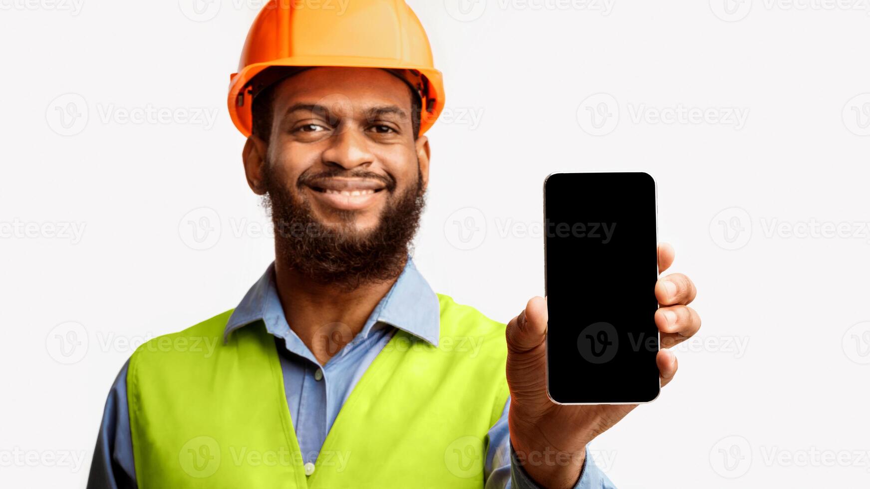 Black Builder Workman Showing Smartphone Blank Screen Standing Over White Studio Background. Panorama, Mockup, Selective Focus photo