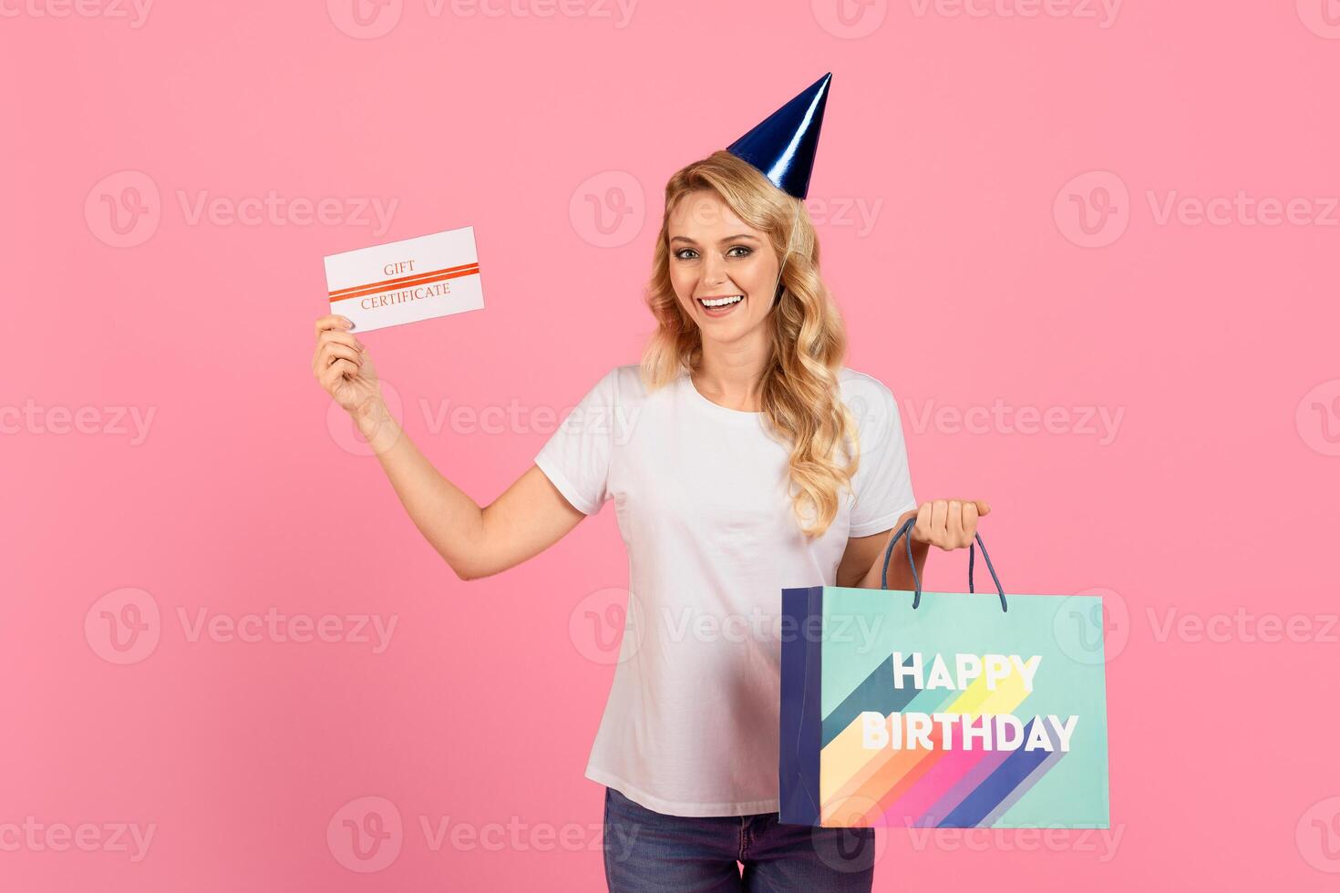 A cheerful young woman wearing a party hat holds a gift certificate and a colorful birthday bag. The backdrop is bright pink, enhancing the festive atmosphere of the celebration. photo