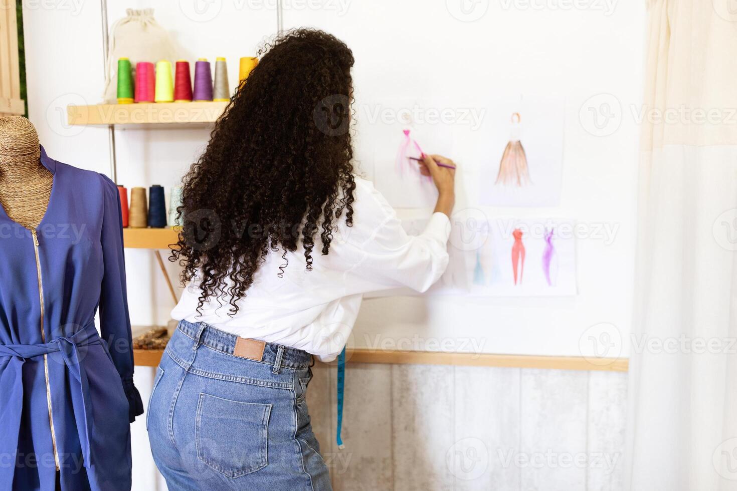 A mature Hispanic woman focused on her design work in an organized office. Surrounded by colorful spools of thread, she sketches fashion ideas on the wall. photo