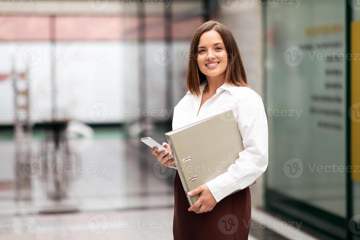 A confident businesswoman stands in a sleek corporate office holding a binder and smartphone. She appears ready to engage in work, showcasing professionalism and style in a modern setting. photo
