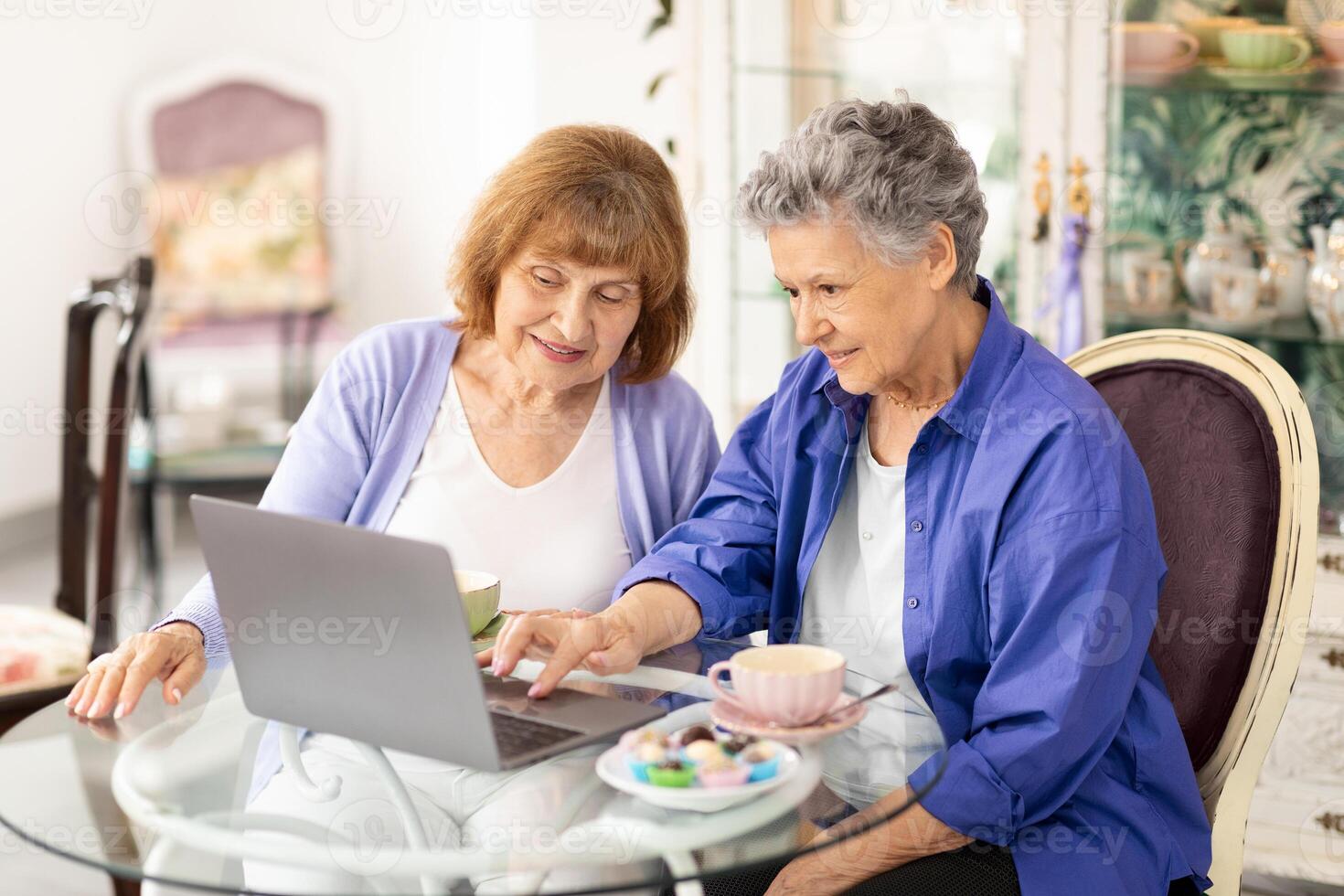 Two elderly women share a relaxing moment at a cafe, engaged in lively conversation while browsing on a laptop. They enjoy tea and colorful pastries, embodying a joyful lifestyle. photo