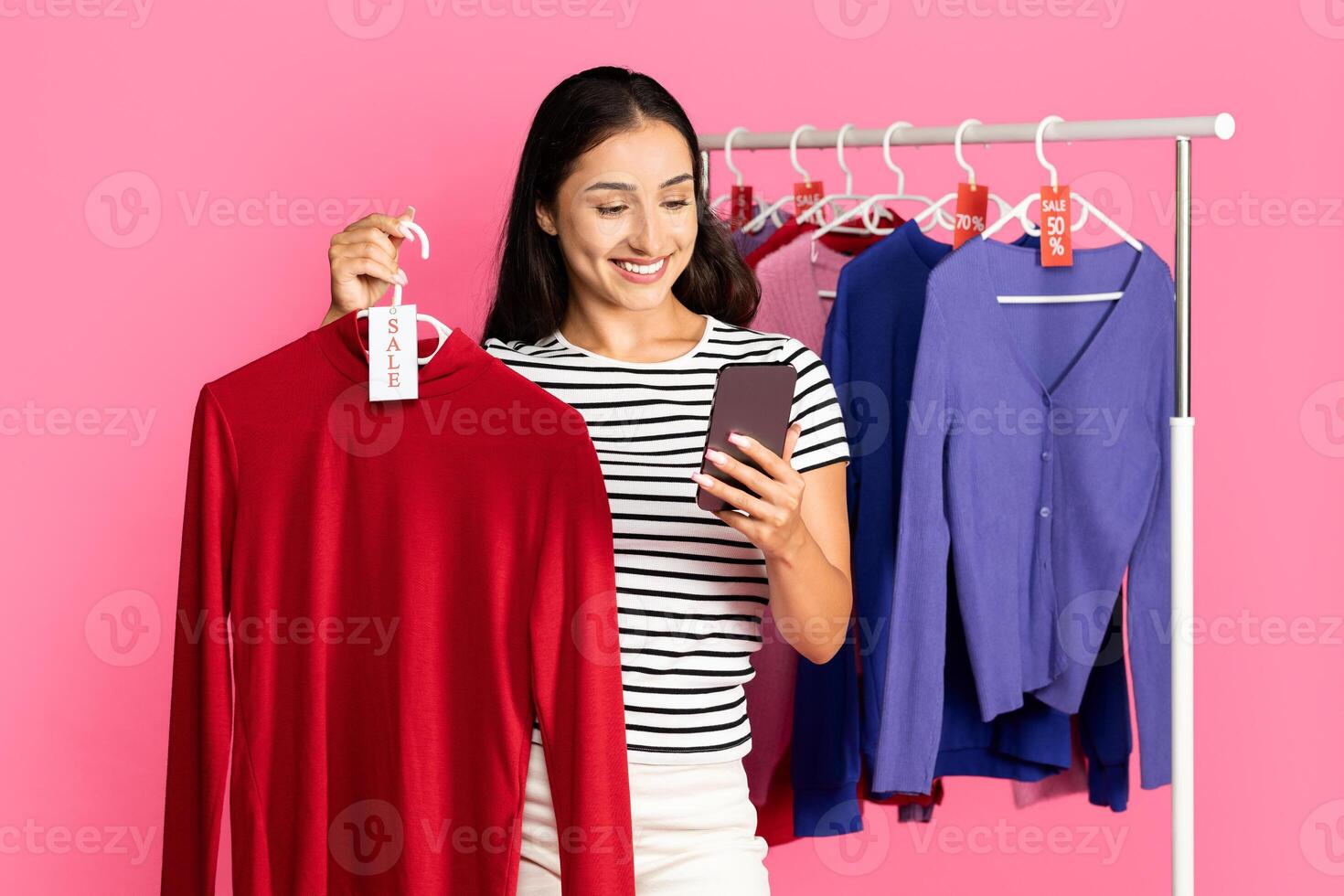 A young woman stands in front of a clothing rack, holding a red sweater with a sale tag. She smiles while looking at her phone, engaging in shopping on a vibrant pink backdrop. photo