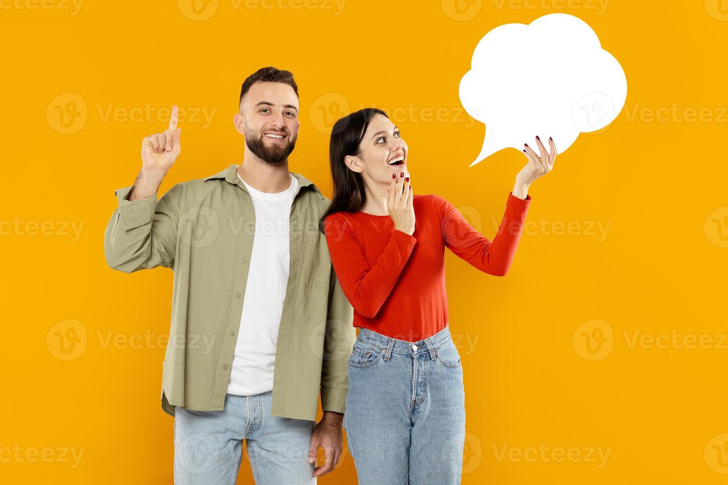 A young couple stands together with expressions of excitement. The man gestures with his finger raised while the woman covers her mouth in surprise, set against a vibrant yellow background. photo