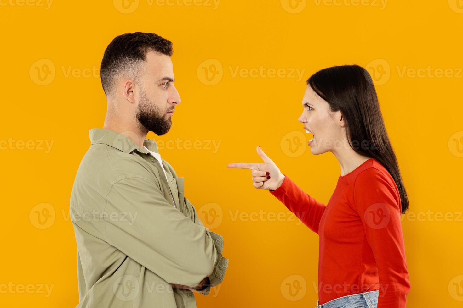 A young couple stands facing each other in a tense moment. The woman appears upset, pointing her finger at the man, who has a serious expression while crossing his arms. photo