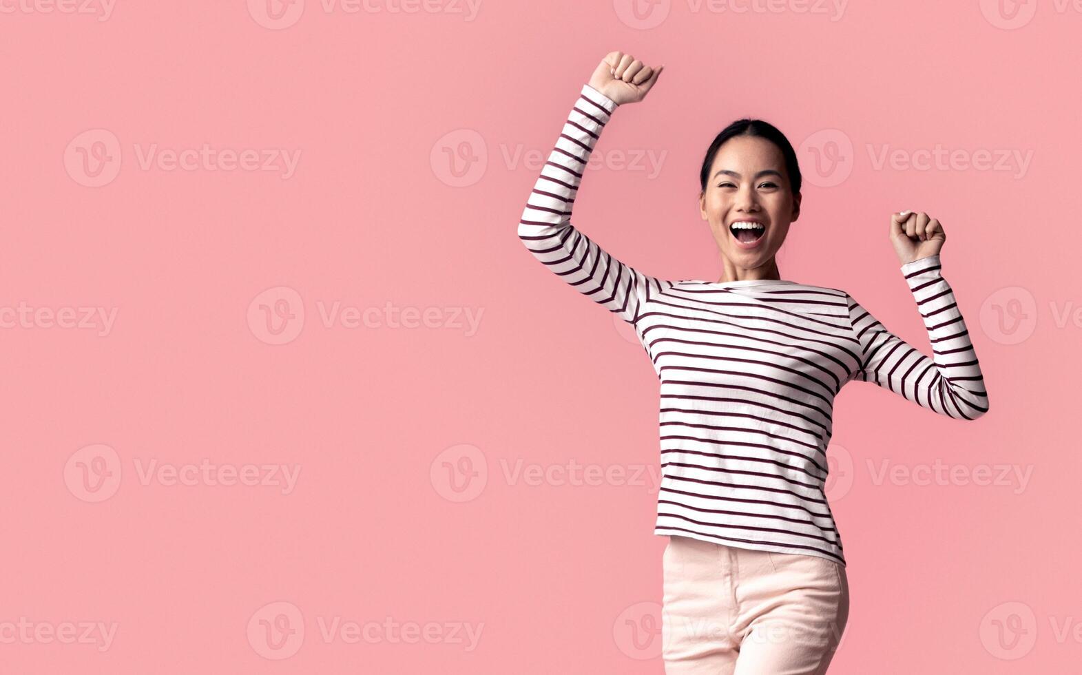 Euphoric Young Asian Woman Jumping With Raised Hands Over Pink Background, Full-Length Shot Of Cheerful Excited Korean Female Celebrating Win With Clenched Fists, Exclaiming With Joy, Copy Space photo