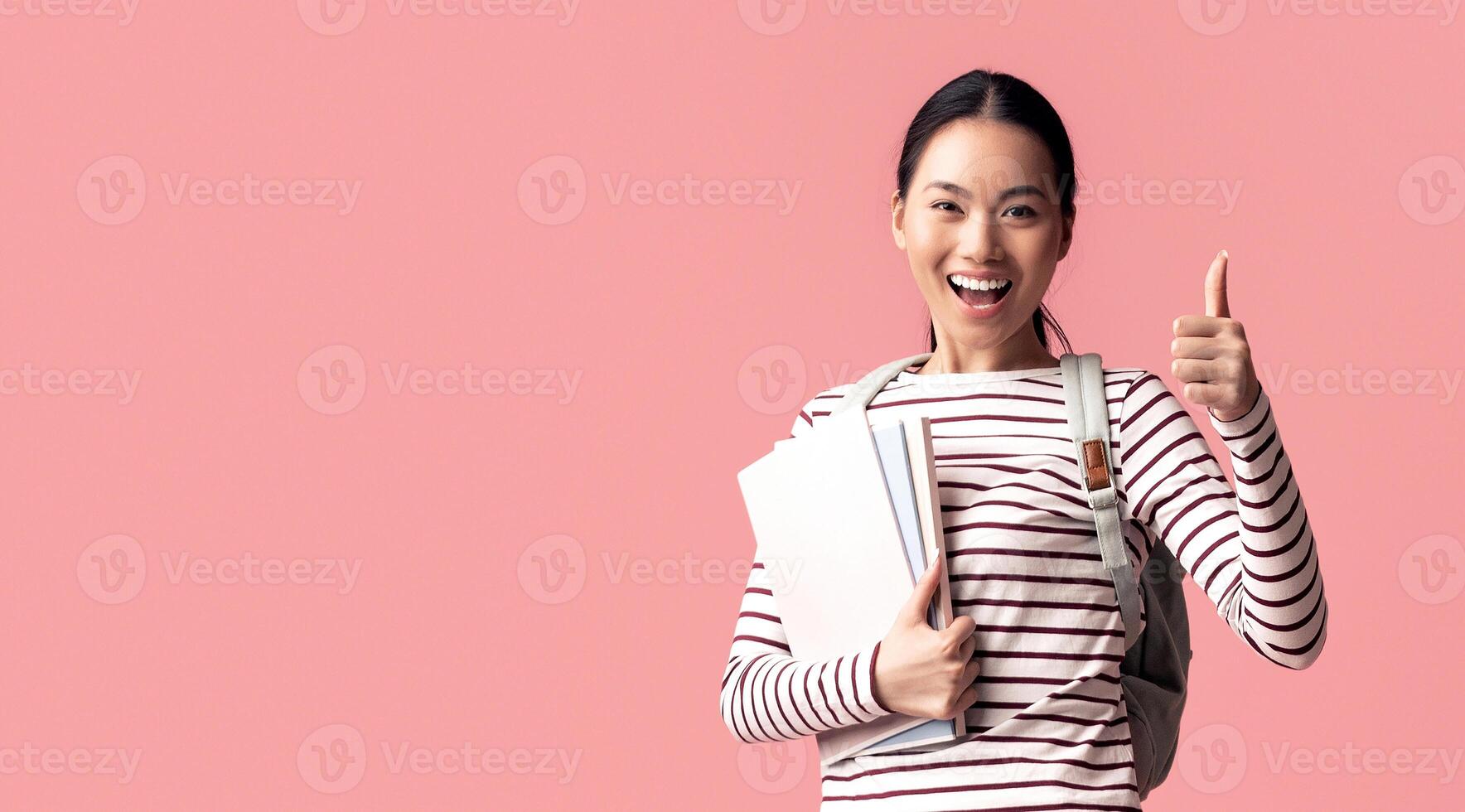 Cheerful Asian Female Student Jumping In Air With Workbooks And Showing Thumb Up, Happy Excited Korean Female Wearing Backpack And Smiling At Camera While Posing Over Pink Background, Copy Space photo