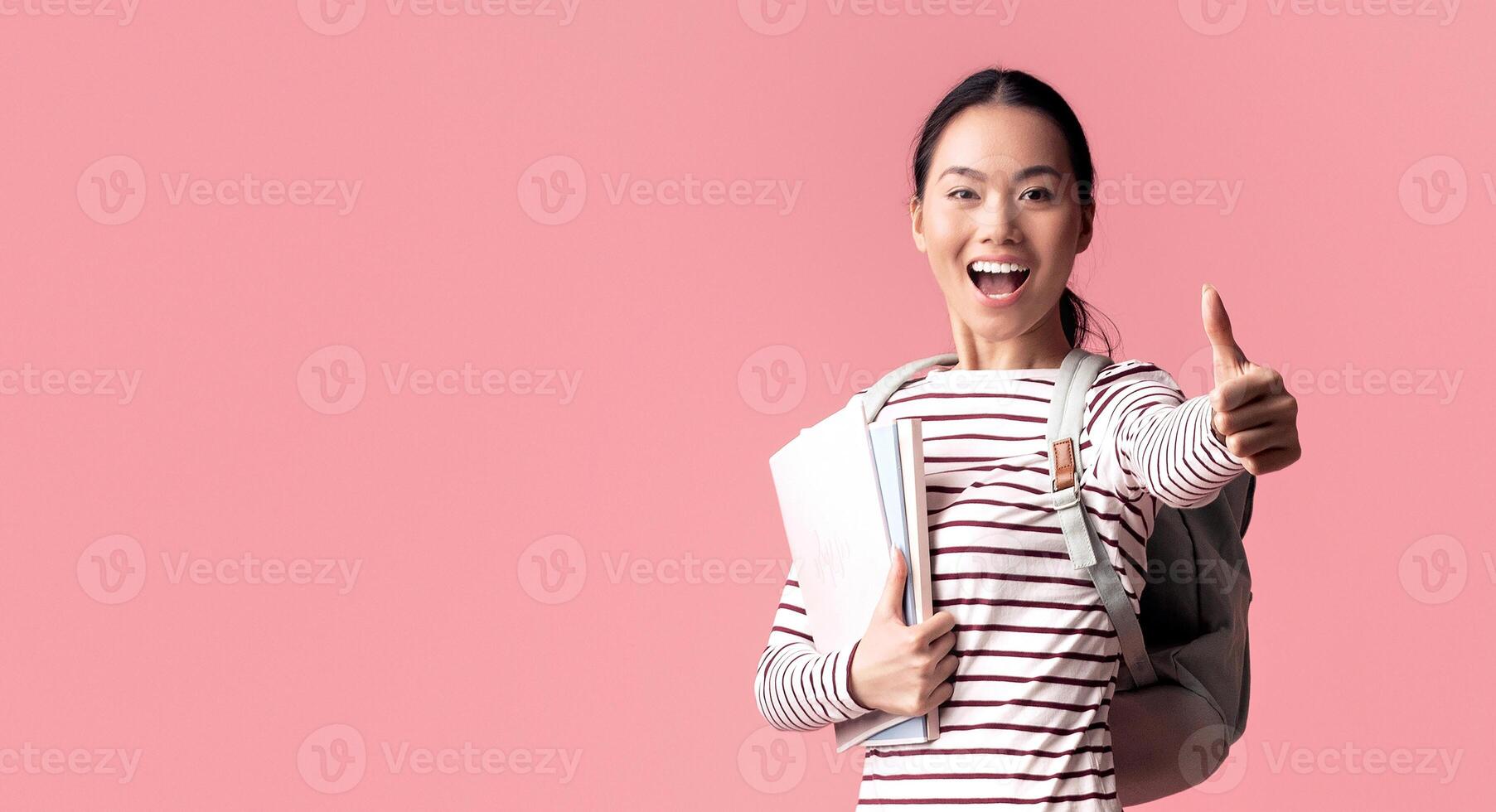 Portrait Of Happy Young Asian Female Student Jumping In Air And Showing Thumb Up At Camera, Cheerful Korean Female Holding Workbooks And Wearing Backpack, Having Fun Over Pink Background, Copy Space photo