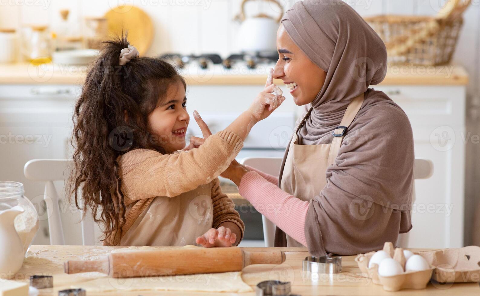 Mother's Day Concept. Cute Little Girl Having Fun With Her Muslim Mom In Kitchen, Playfully Touching Noses Of Each Other While Preparing Dough For Cookies, Enjoying Spending Time Together, Closeup photo