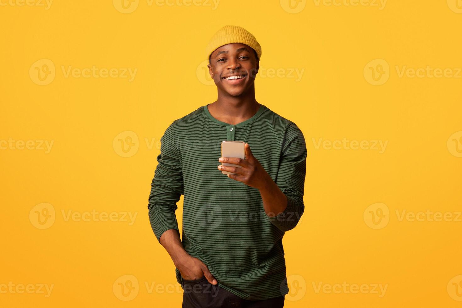 Online Communication. Portrait of cheerful young black man holding smartphone and smiling at camera, happy millennial african guy using cellphone while standing over yellow background, copy space photo
