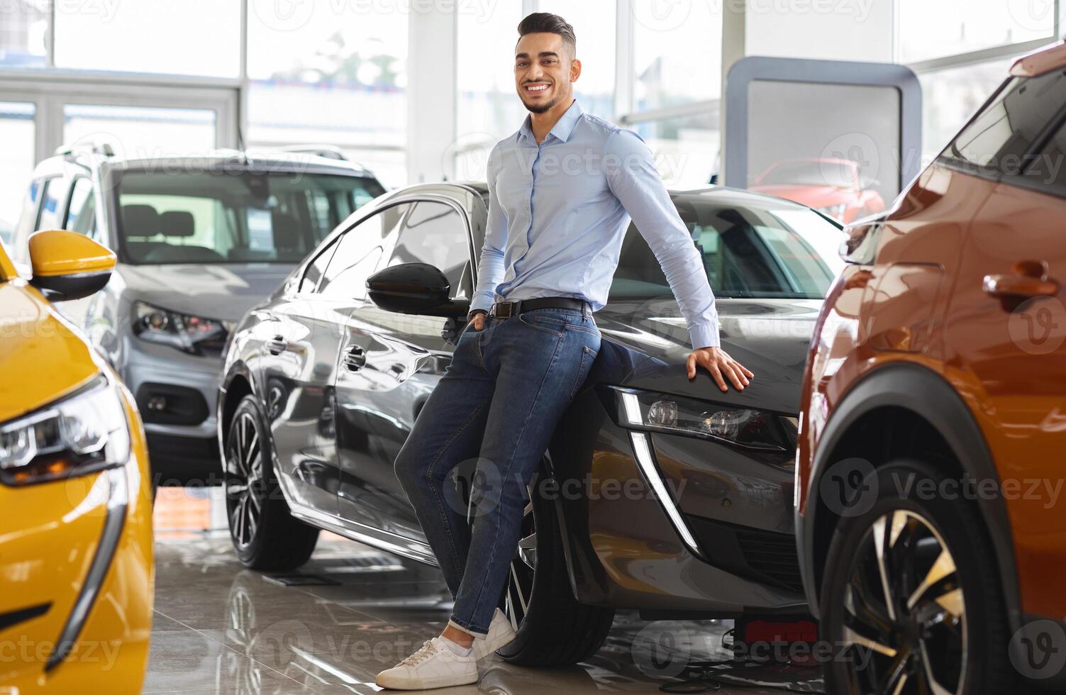 Confident wealthy middle eastern businessman buying new car in luxury auto showroom, cheerful arab guy standing by nice black sports automobile and smiling at camera, copy space, full length photo