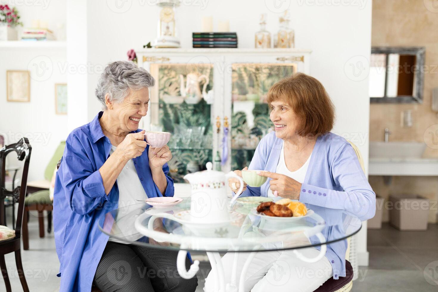 Two elderly women relax in a bright cafe, sharing laughter and sipping tea. They savor pastries while enjoying each other's company during retirement. photo