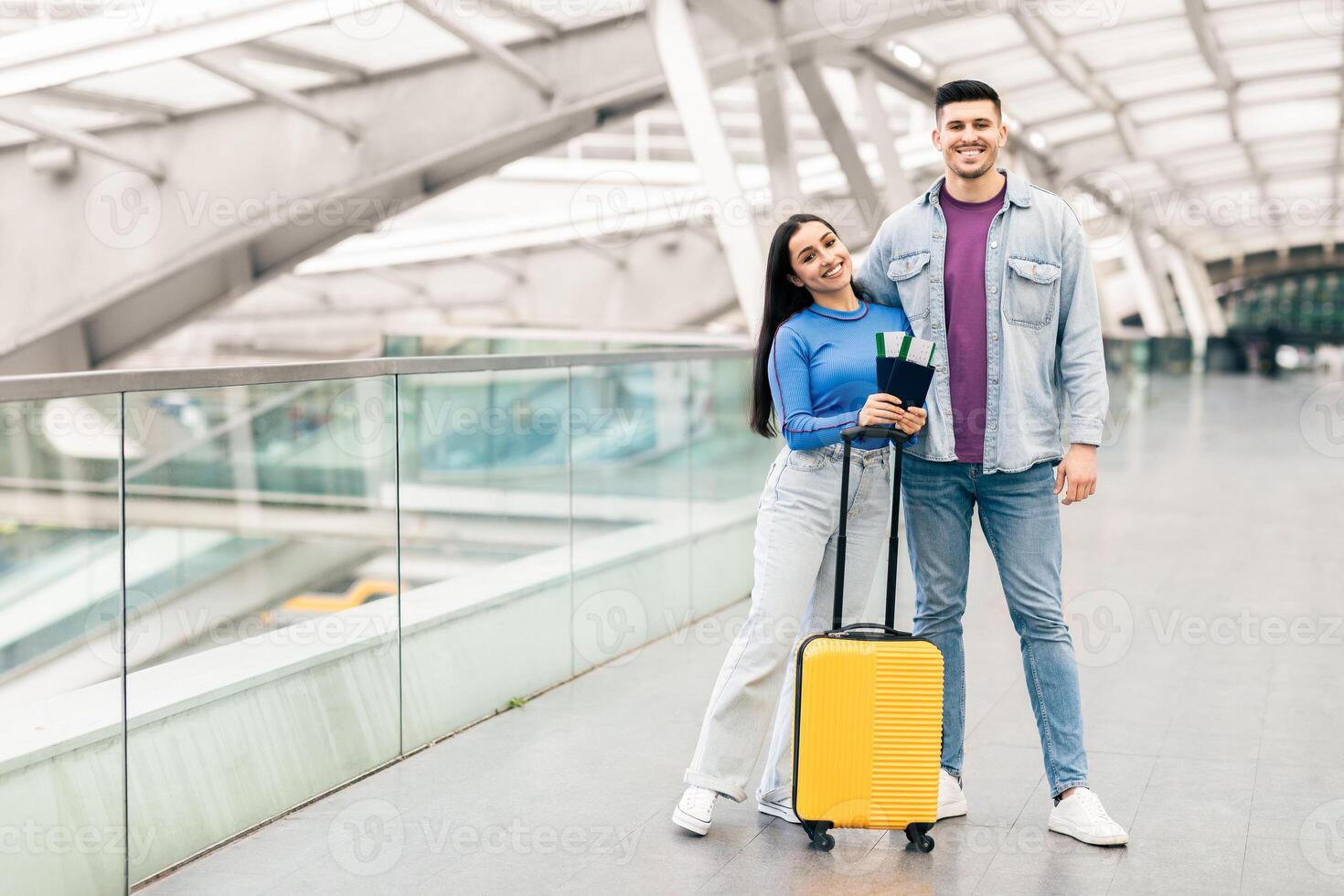 A young couple stands at the airport with smiles, ready to embark on their travel adventure. They hold tickets and are accompanied by a yellow suitcase, showcasing their joy for the trip ahead. photo