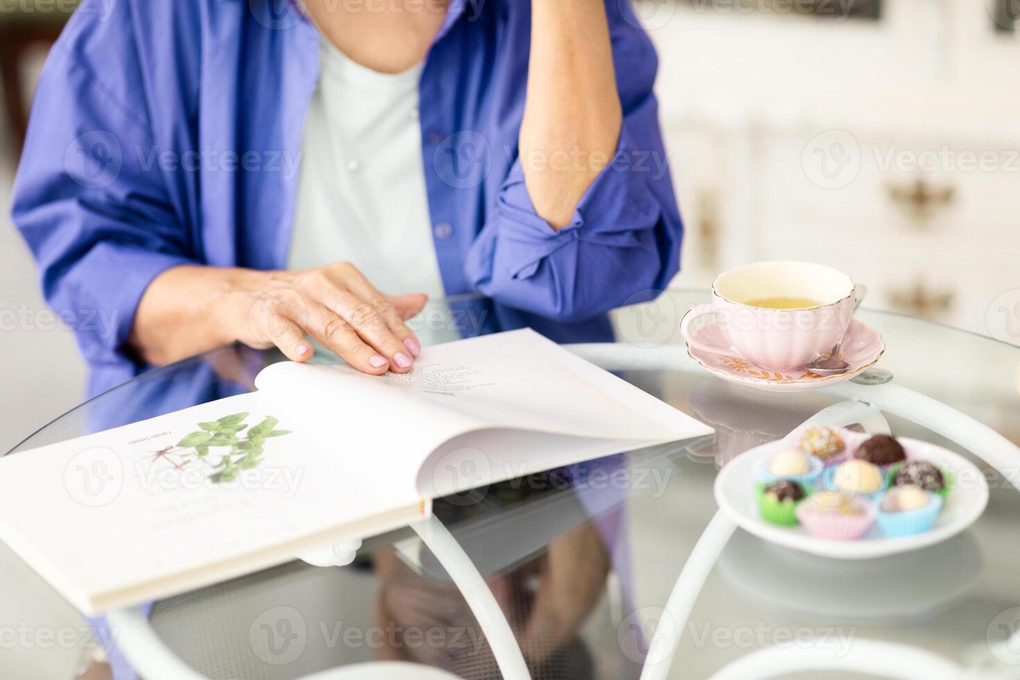 A retired woman in casual attire sits at a cafe table, reading a book while sipping tea. Colorful candies are placed beside her, creating a warm and inviting atmosphere. photo