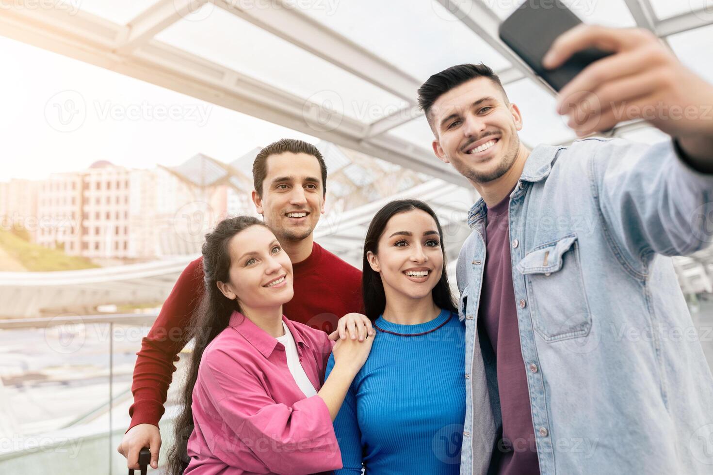 A young couple, along with two friends, happily poses for a selfie indoors at the airport, capturing memories before their weekend getaway. The atmosphere is filled with excitement. photo