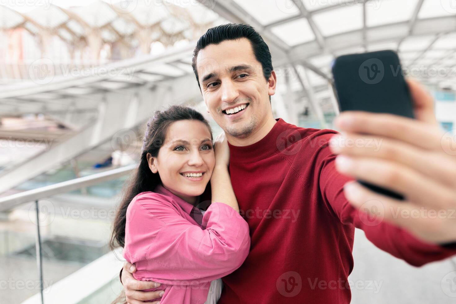 A young couple is happily taking a selfie at the airport. They are embracing each other while showcasing their excitement for their upcoming travel adventure. photo