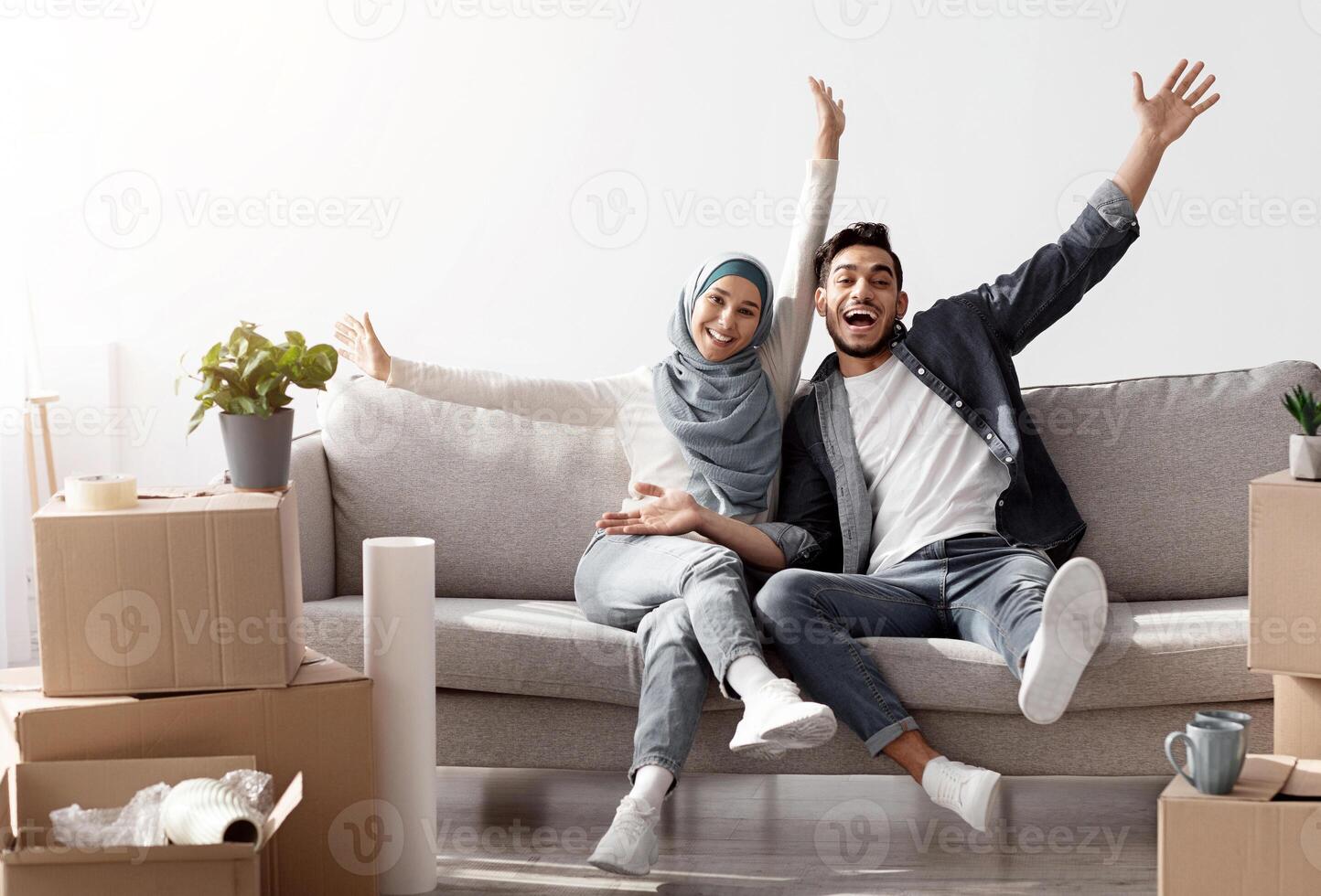 Emotional muslim family celebrating moving to their new house, sitting on new couch among paper boxes with stuff and having break while unpacking belongings, raising hands up and smiling, copy space photo