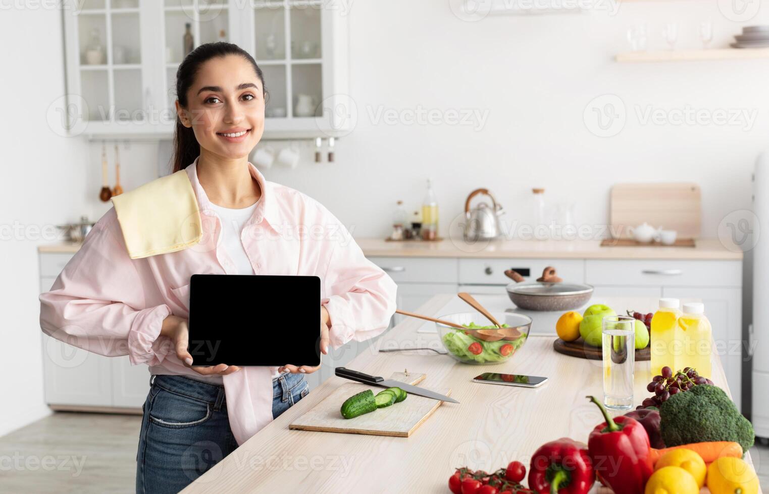 Cheerful Young Lady Showing Empty Digital Tablet Screen Posing Standing In Modern Kitchen At Home. Smiling Woman Recommending Cooking Application Or Website With Healthy Online Recipes. Mock up photo