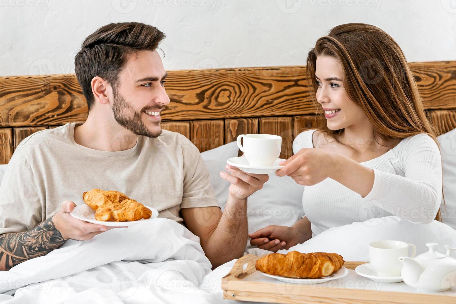 Romantic breakfast of couple in bed. Smiling attractive young woman holding tray with croissant and giving a cup of coffee to man with beard, free space photo
