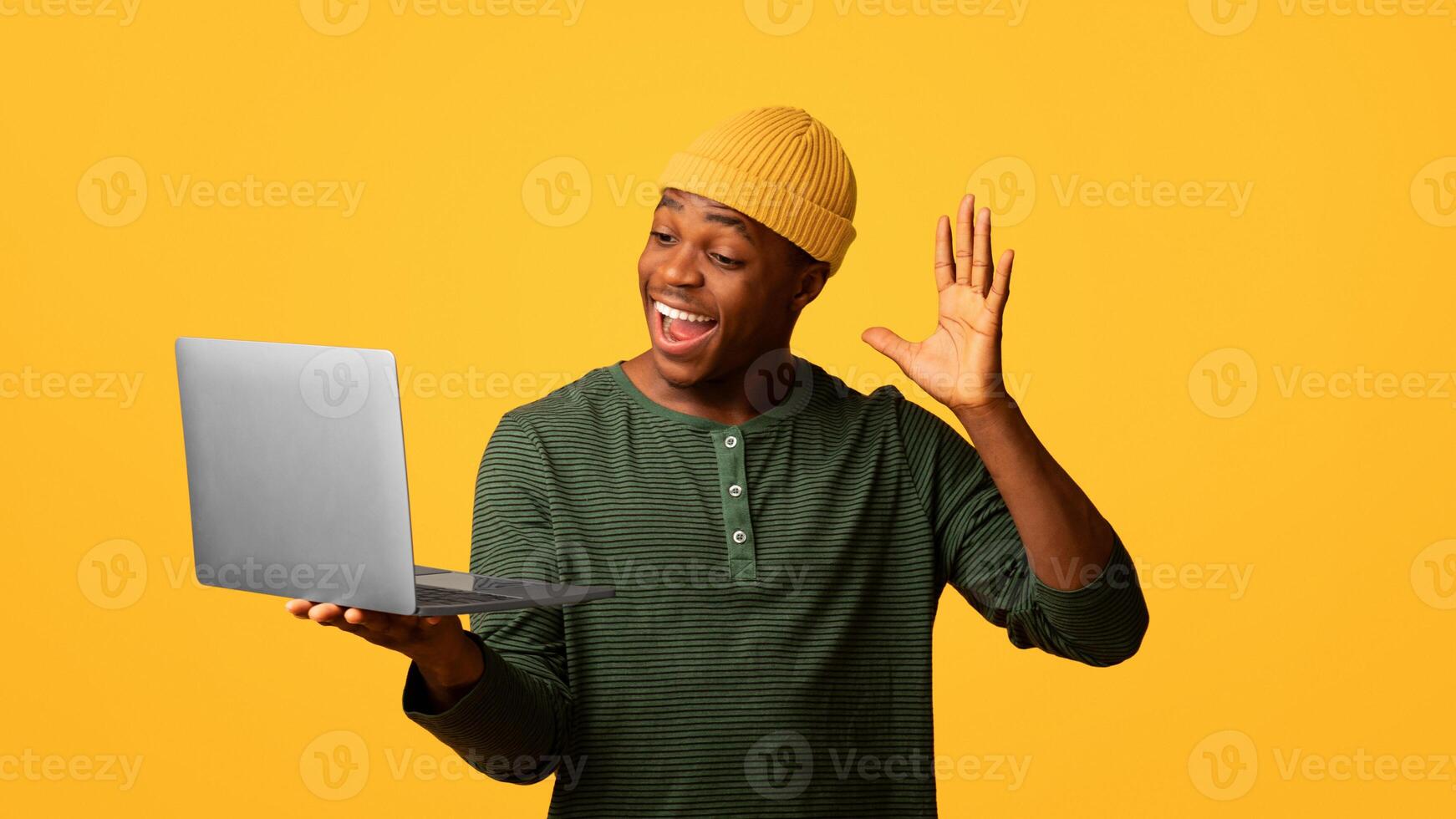 Cheerful african american guy making call with laptop, waving hand at web camera, friendly young black man greeting somebody while standing on yellow background, enjoying online communication photo
