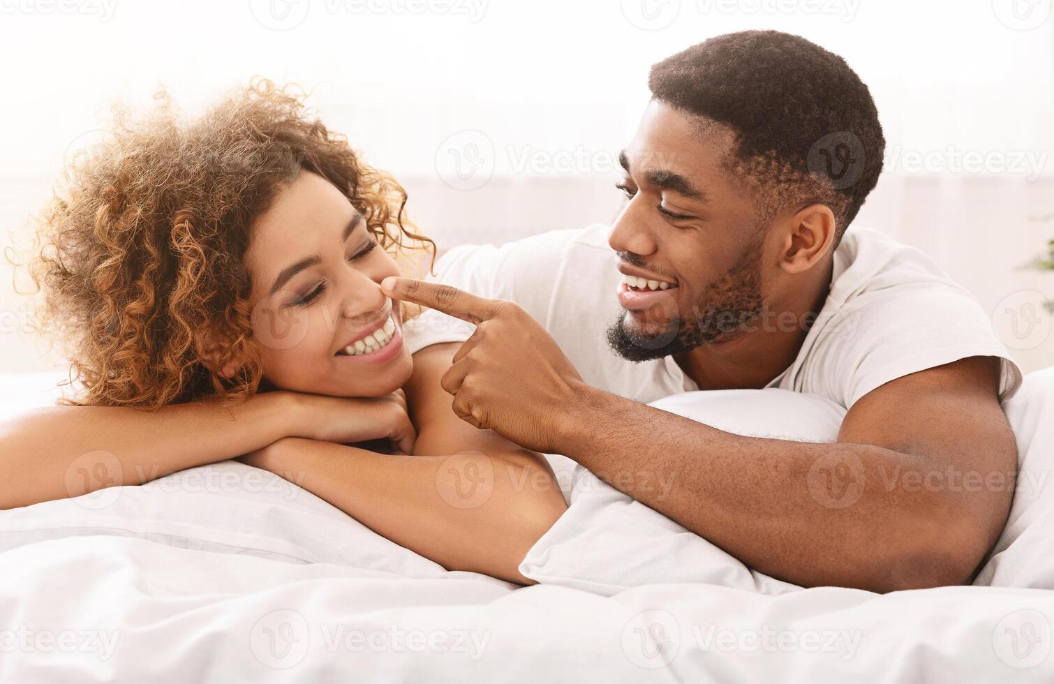 Couple romance. Playful african man flirting with wife, touching her nose in bed photo