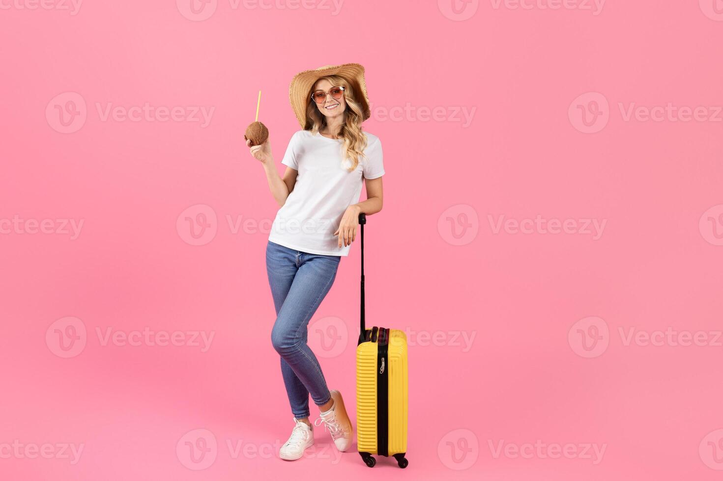 A young woman stands confidently next to a bright yellow suitcase holding a coconut drink. She wears sunglasses and a straw hat, ready for a fun summer getaway in a pastel pink setting. photo