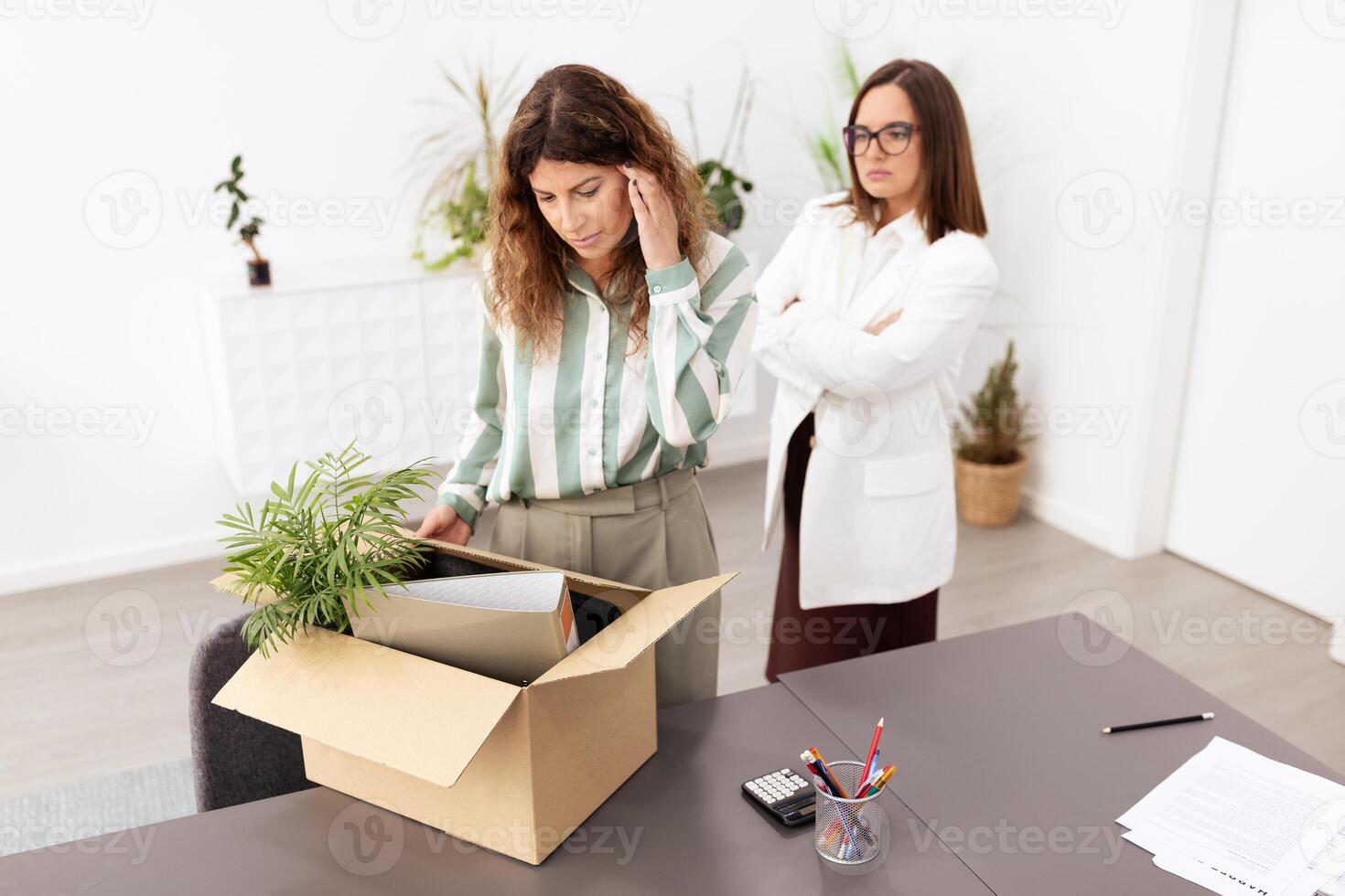 Two women in a modern office setting handle a box of documents. One woman appears concerned while the other observes, highlighting the dynamics of corporate responsibilities. photo