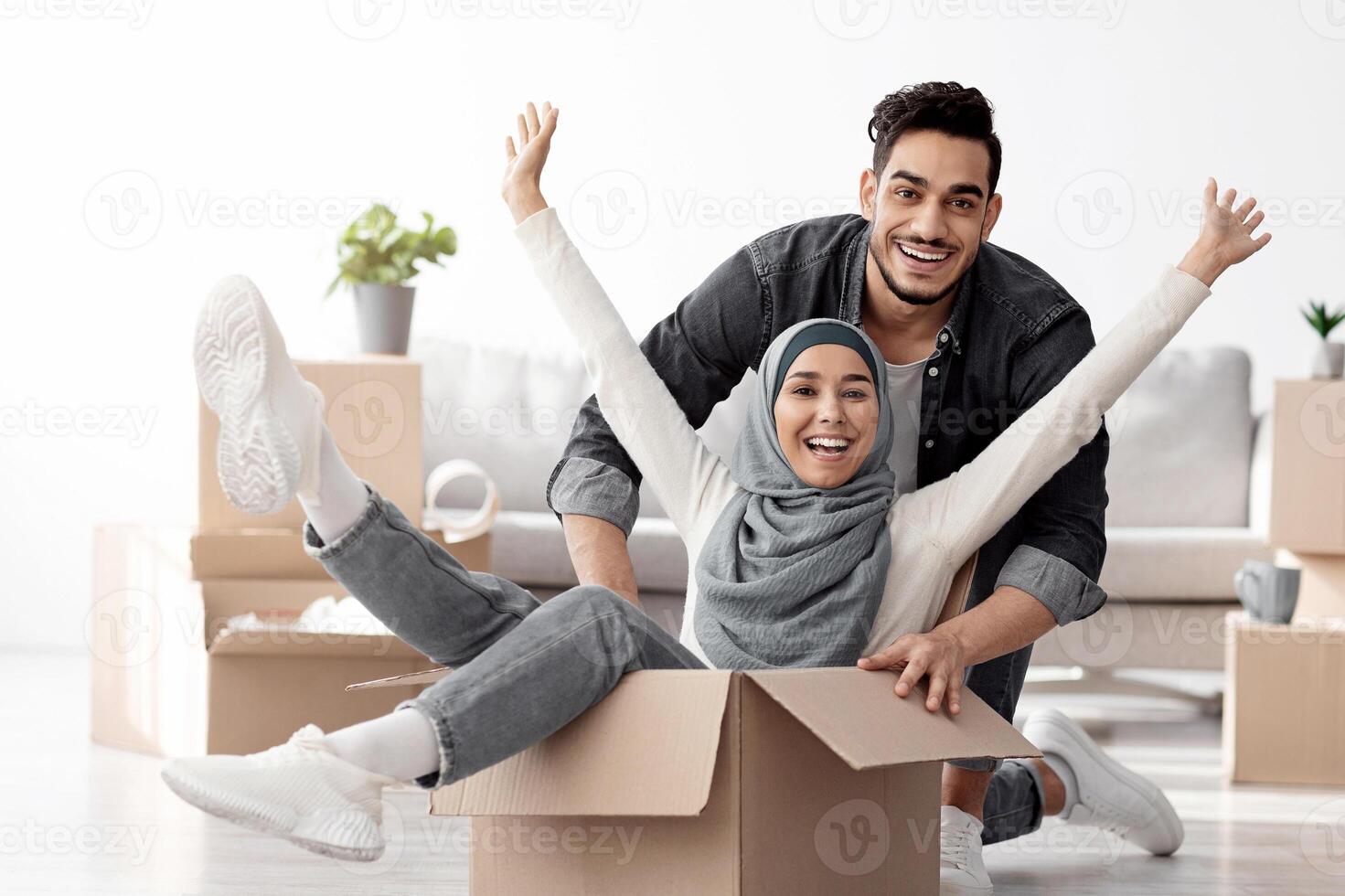 Positive muslim family playing with packing supplies while moving to their new house. Happy lady in hijab sitting in paper box and raising hands up, having fun with handsome husband while unpacking photo