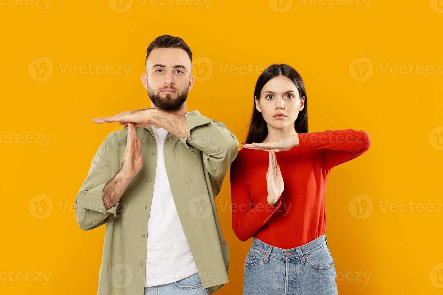 A young Caucasian couple stands together, playfully using hand signals to convey affection. Their vibrant expressions reflect a strong bond, set against a bold, bright yellow backdrop. photo