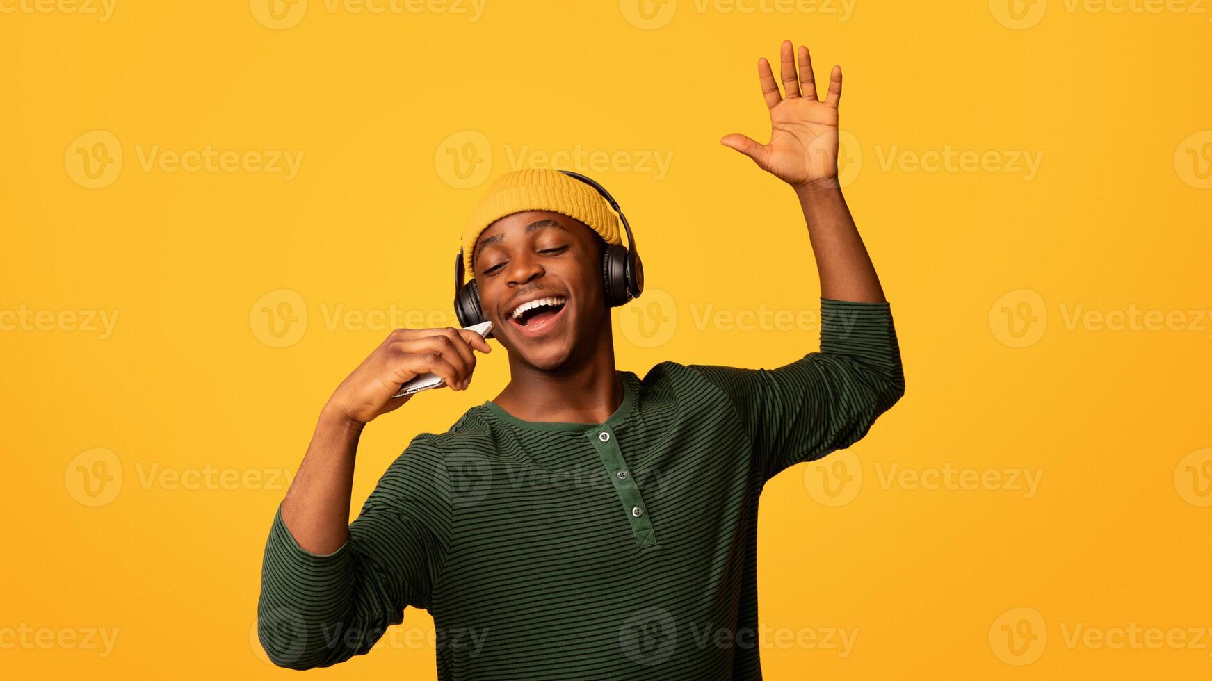 Technologies For Fun. Cheerful black hipster guy listening music in wireless headphones and singing, positive young african man using smartphone as microphone, having fun on yellow background photo