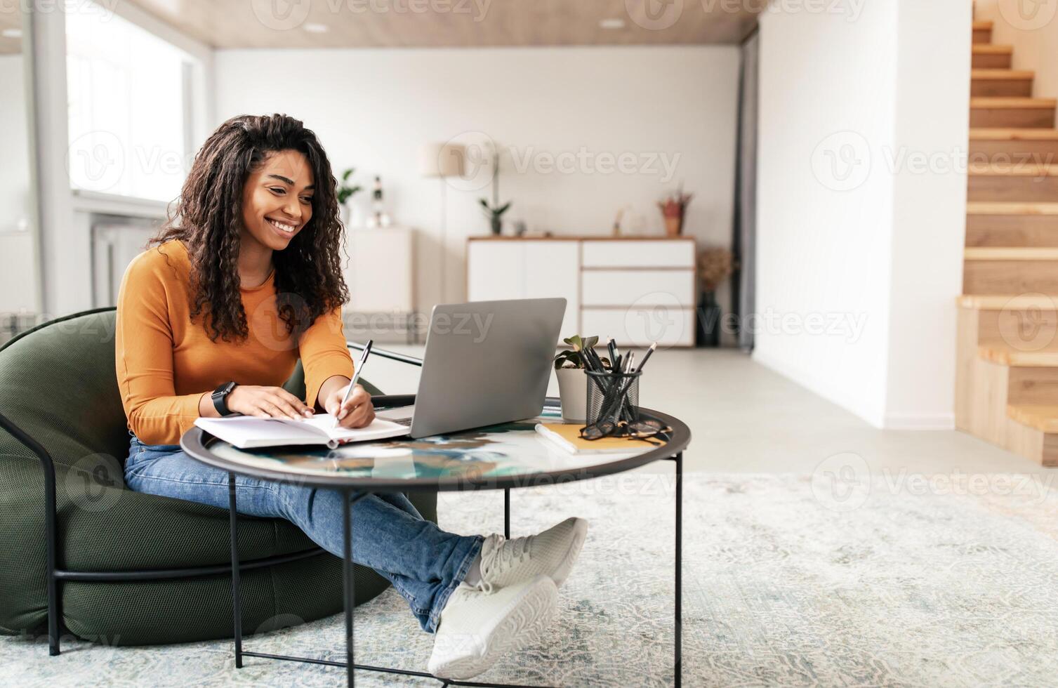 Online Training Concept. Smiling casual black lady sitting at tea coffee table working on pc laptop writing letter in paper notebook, taking notes watching weninar, free copy space, full body length photo