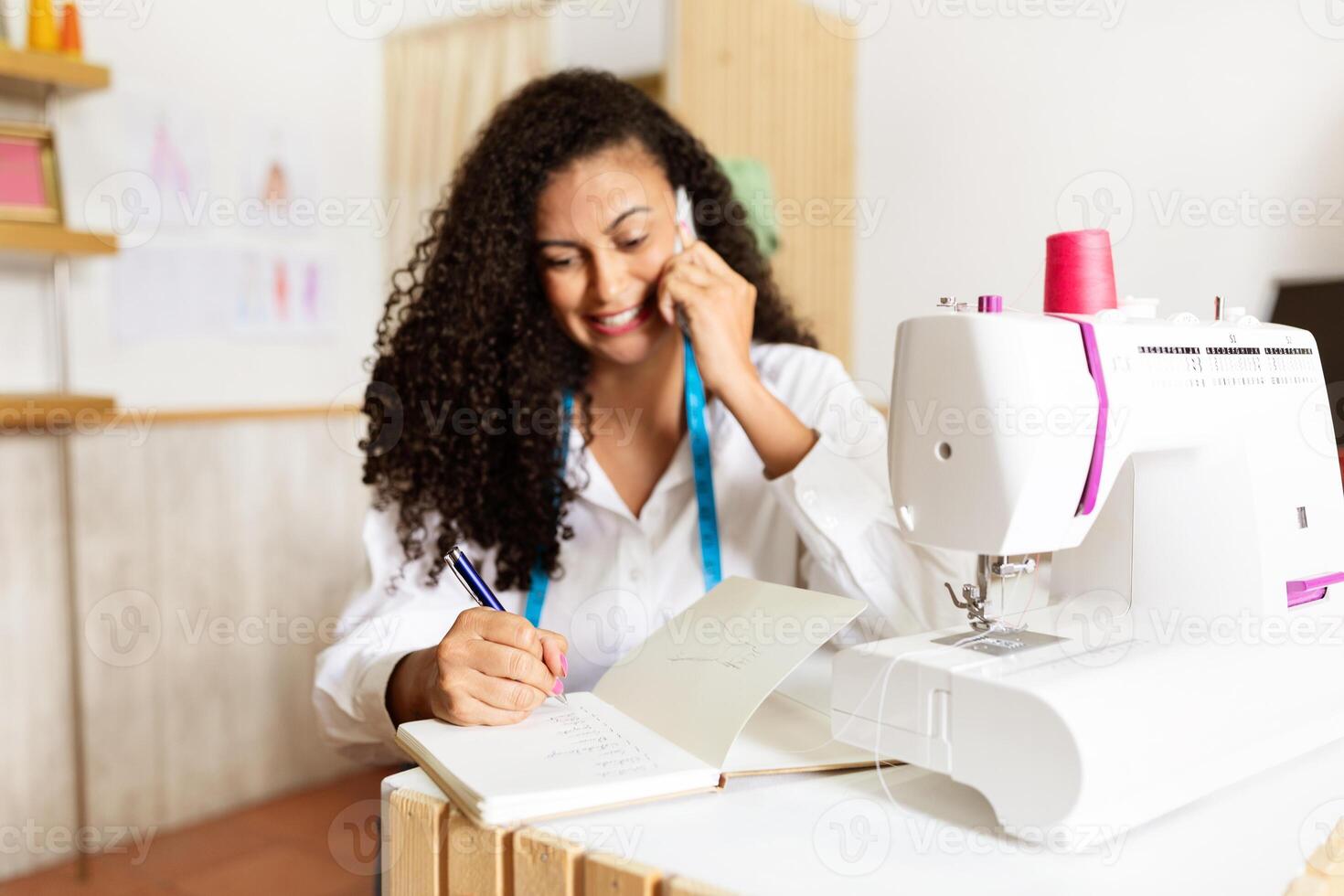 A mature woman with curly hair is taking notes while speaking on the phone in her design studio. She is surrounded by fabric and tools, demonstrating her dedication to her craft. photo