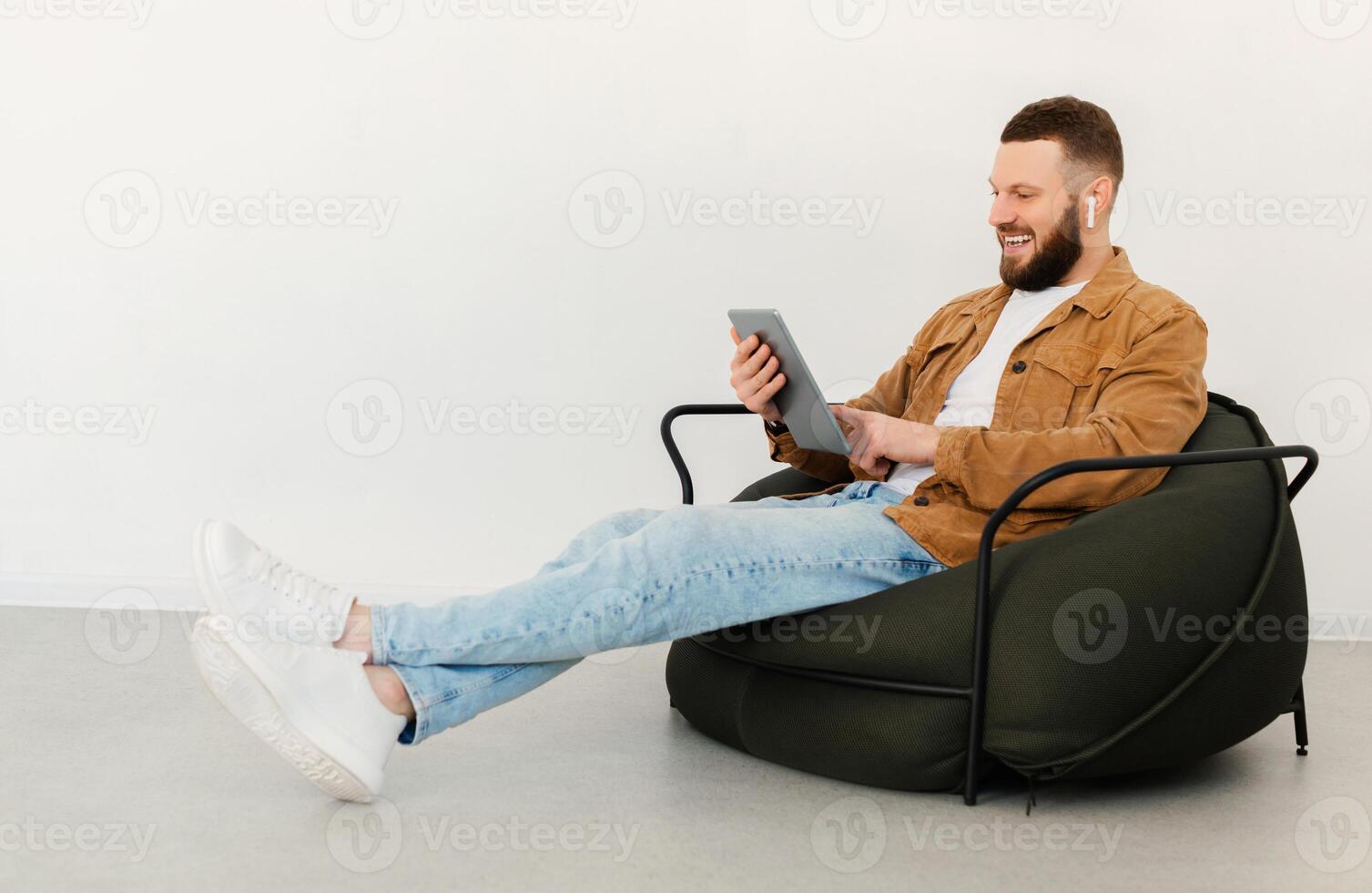Side View Of Man Using Tablet Computer And Earbuds Earphones Watching Movie Online Sitting In Chair Over White Wall Indoors On Weekend. E-Learning, Internet And Technology Concept photo