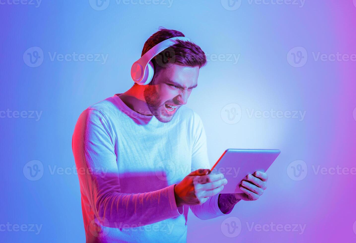 Emotional guy and online games. Young man in big headphones plays in tablet with luminous screen, free space photo