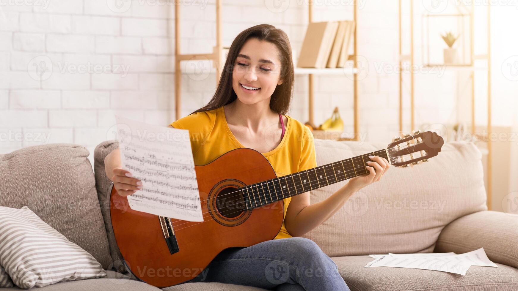 Fun stay home activities. Millennial girl with music sheet and guitar sitting on sofa indoors photo