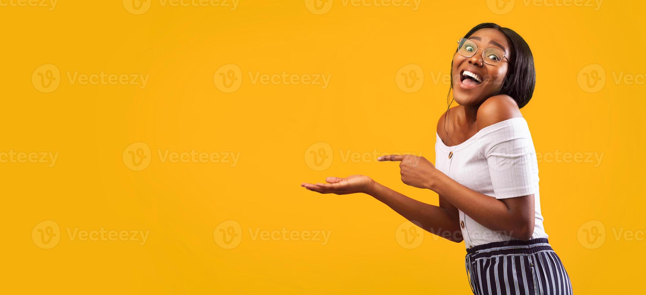 Excited African American Girl Pointing Finger At Open Palm Standing Over Yellow Background. Studio Shot, Panorama With Empty Space photo
