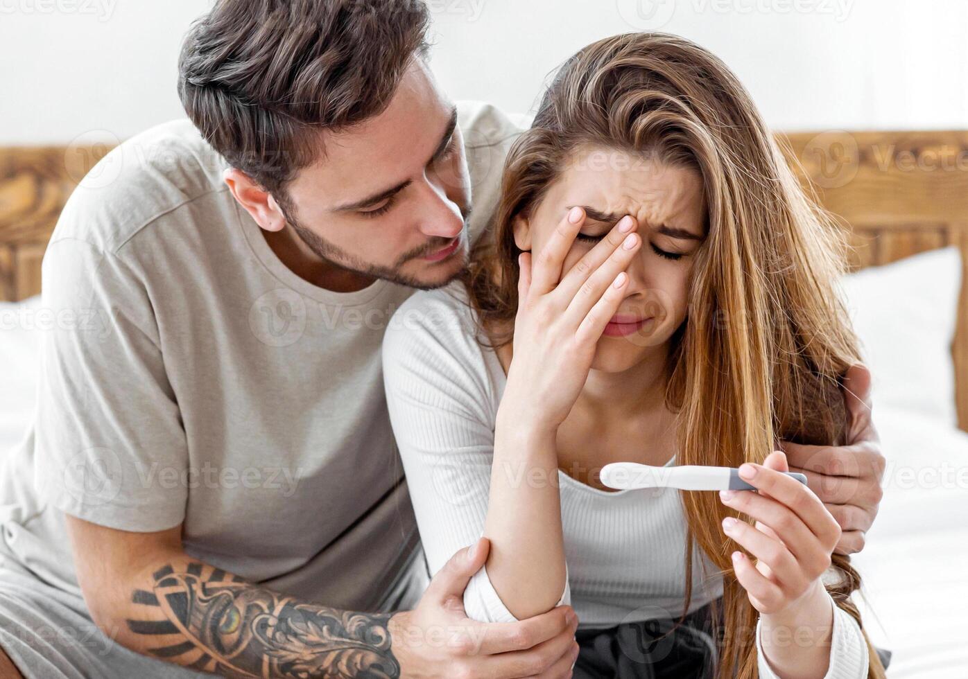 Calm down in difficult times and problems with conception. Sad man hugs crying woman and looks at pregnancy test in bedroom interior photo
