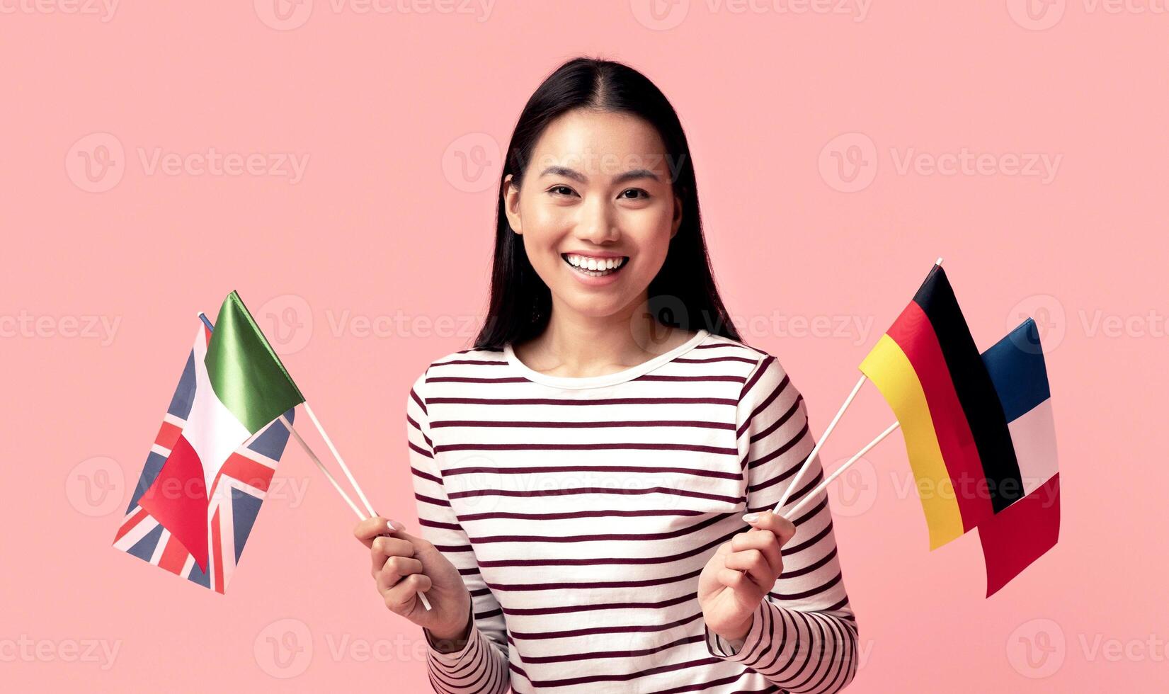 Learn Foreign Languages Online. Happy Asian Woman Holding International Flags And Using Laptop While Sitting On Floor Over Pink Studio Background, Young Korean Student Enjoying Distance Learning photo