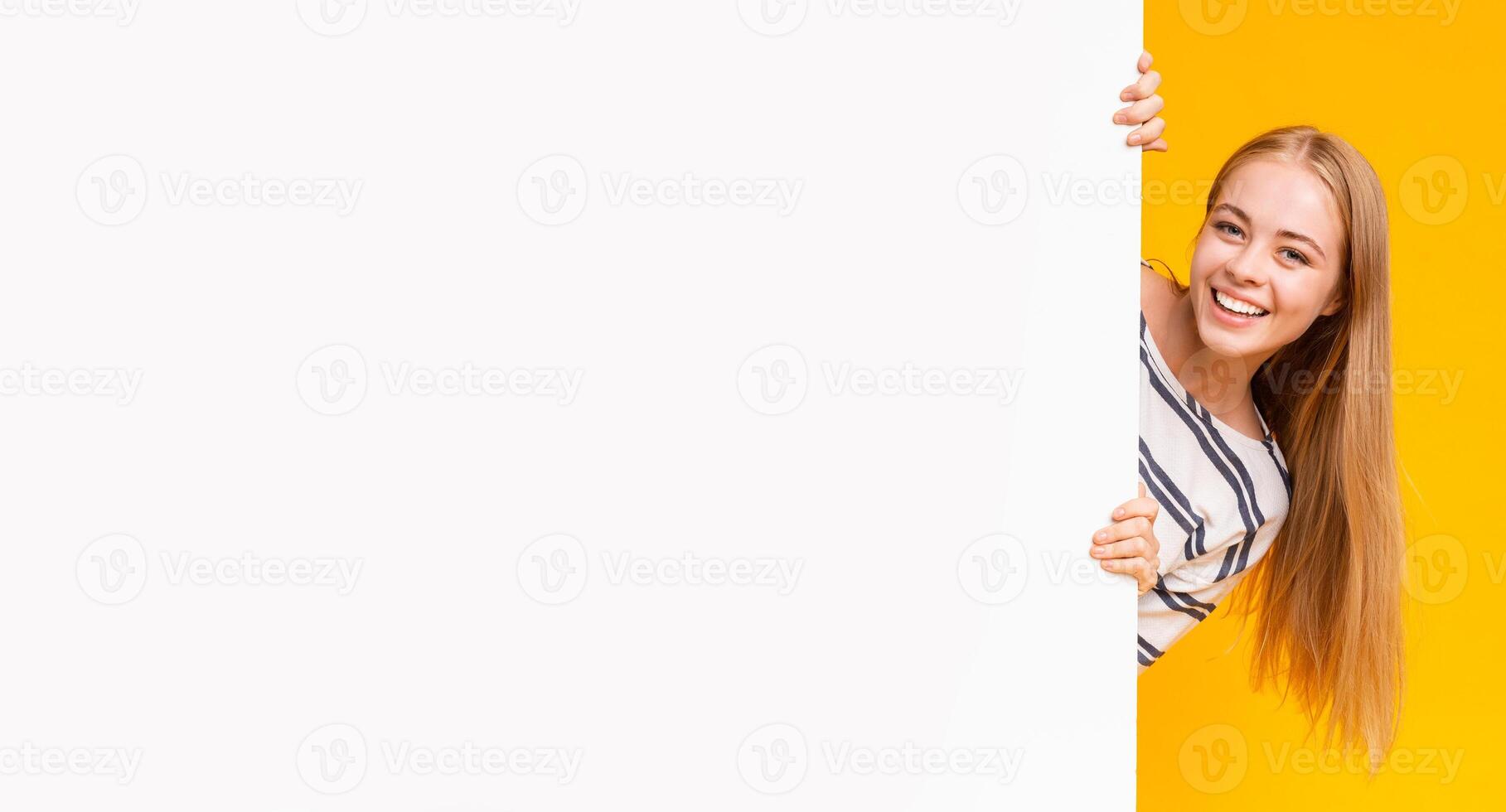 Excited smiling girl looking from behind blank advertising board over yellow background, panorama photo