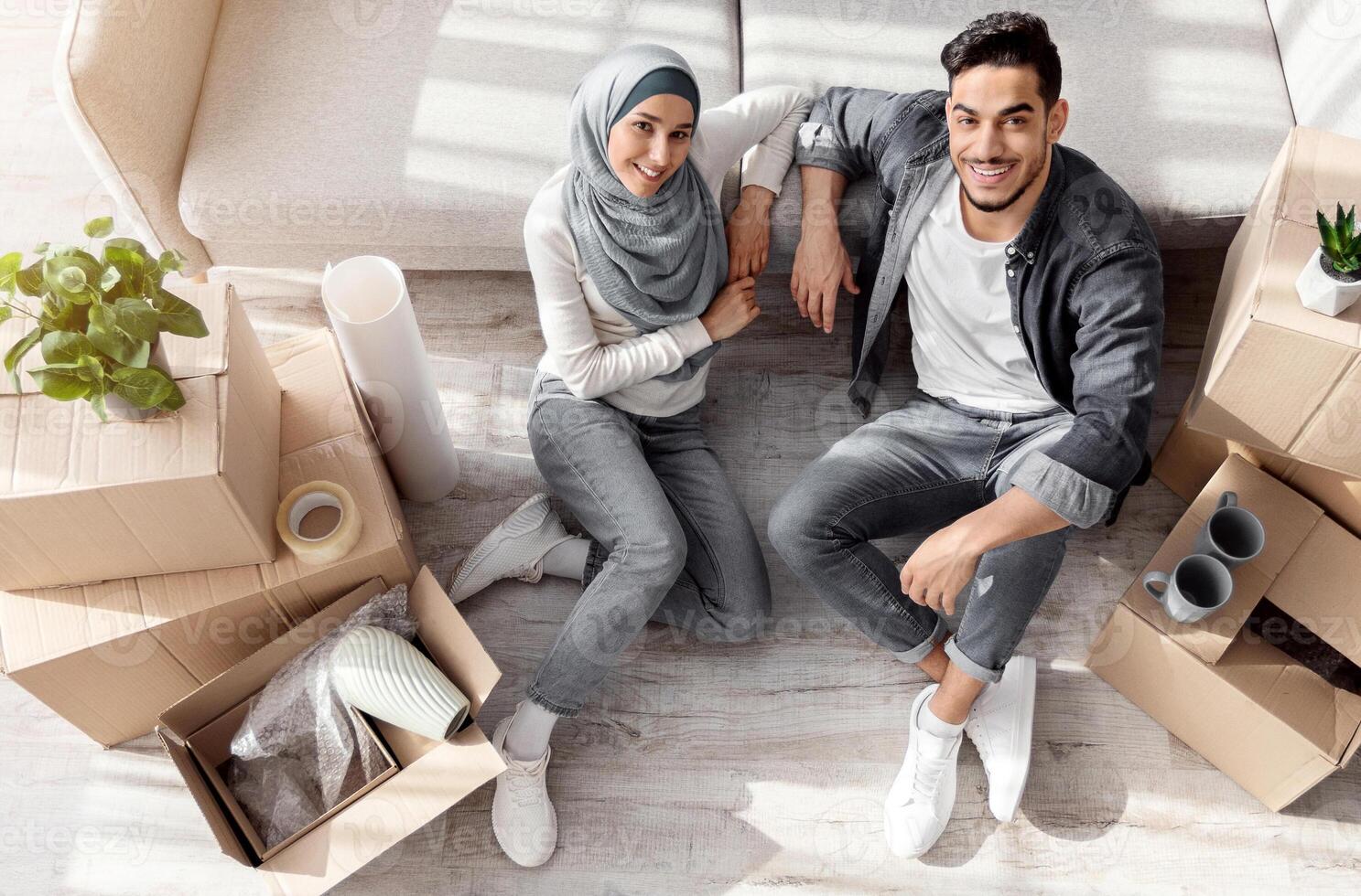 Top view of young muslim family sitting on floor by couch among moving supplies, having break while moving to their new apartment. Happy arab man and woman in hijab with paper boxes photo